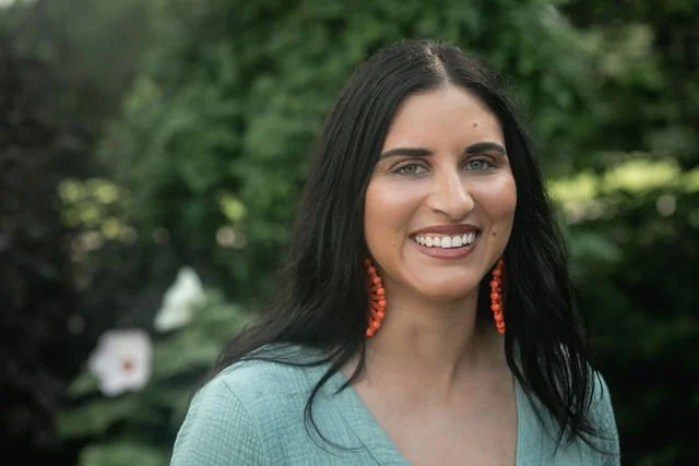 A smiling woman with long dark hair wearing coral earrings and a light blue top standing outdoors with green foliage in the background.