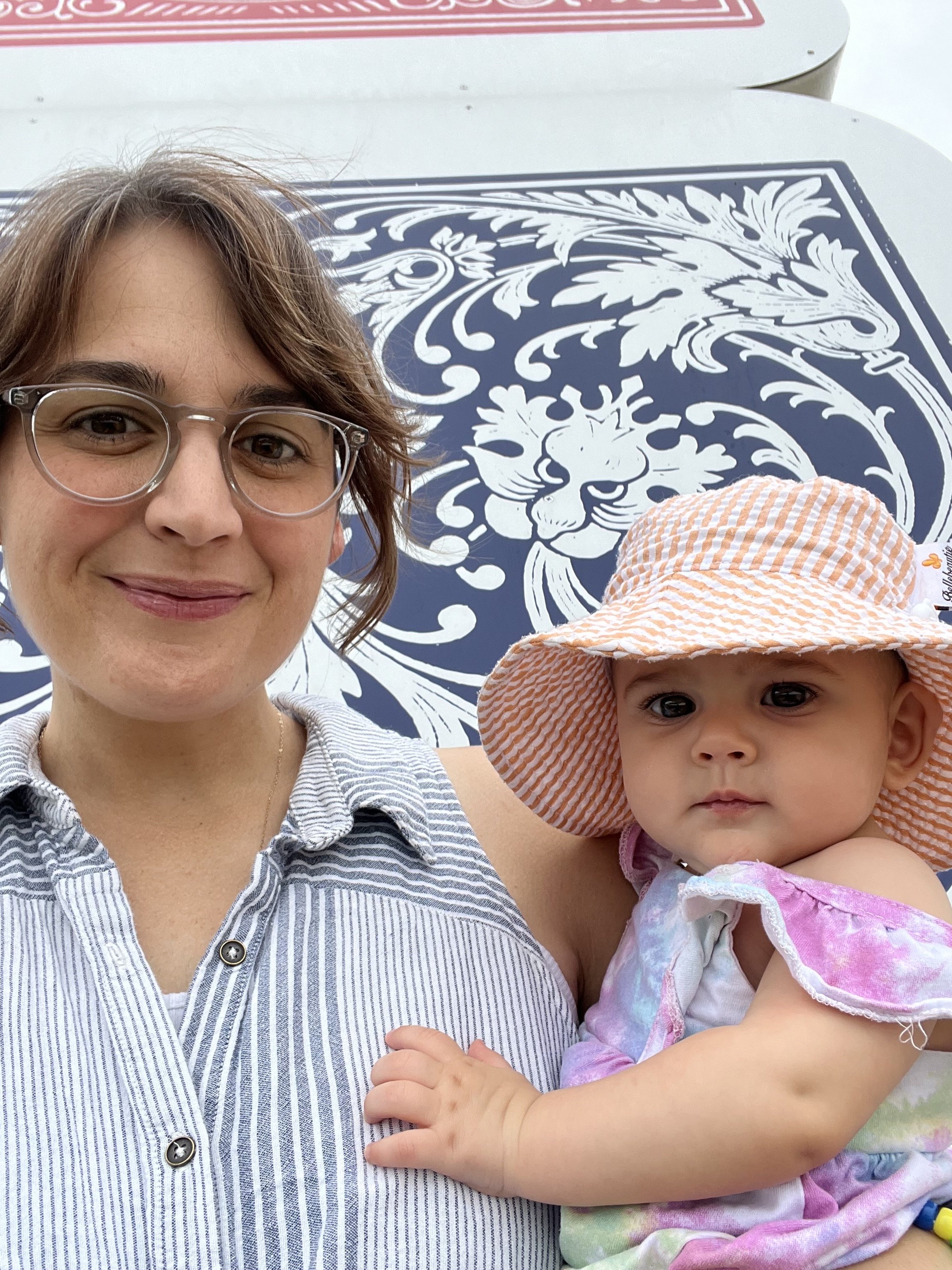 A woman with short, wavy hair and glasses smiling at the camera, holding a young girl in a pink and purple outfit with a colorful sun hat, against a background with a decorative lion and floral design.