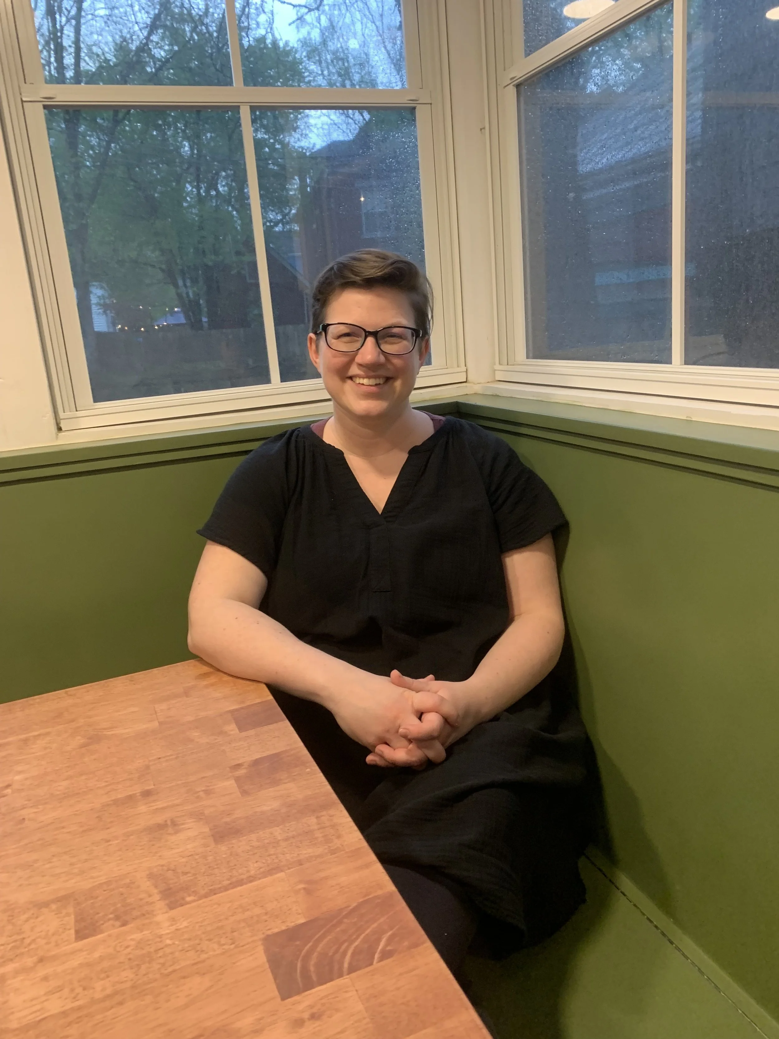 A person with short hair, glasses, and a black shirt sitting at a wooden table near a green wall, with large windows showing trees outside during evening or rainy weather.