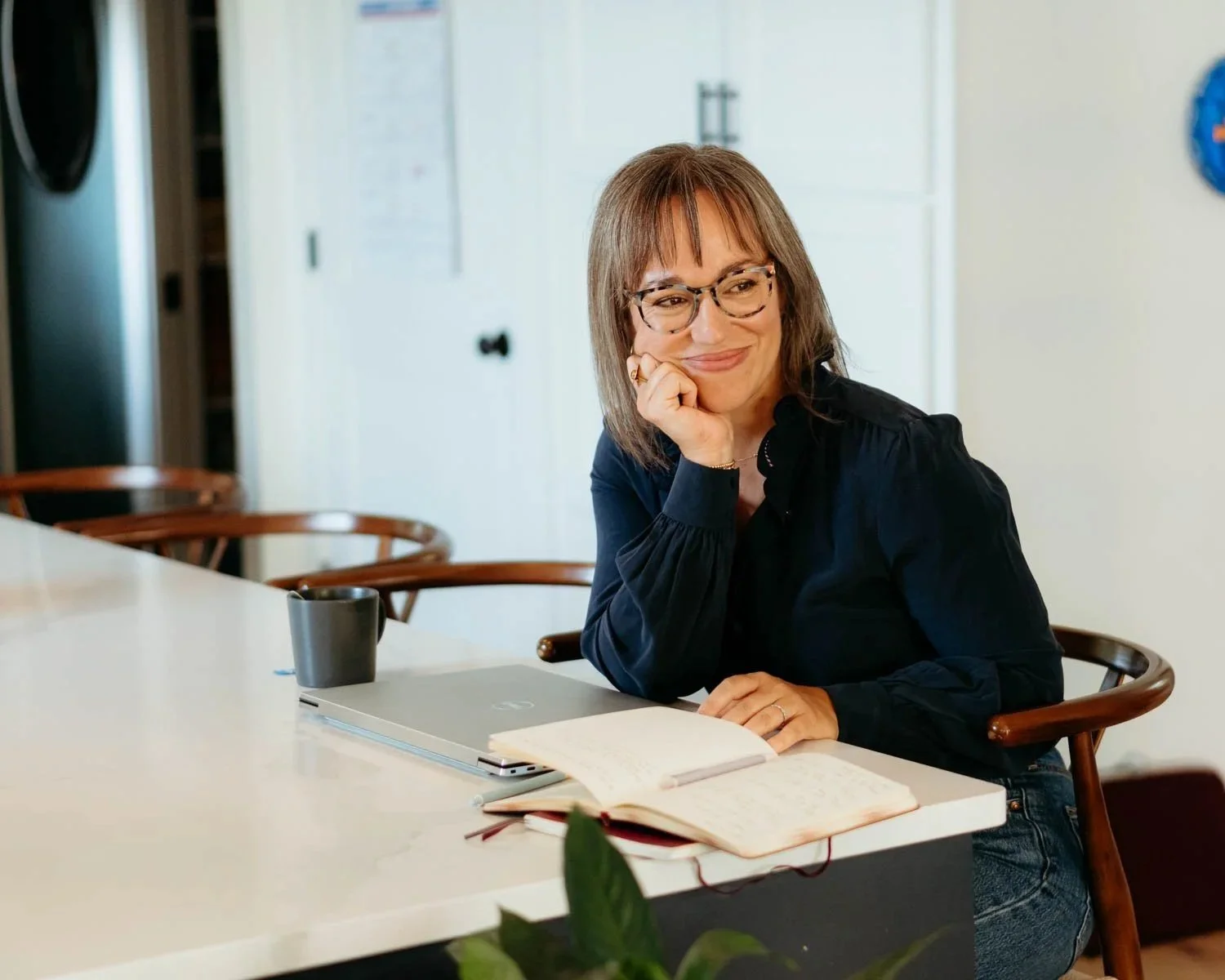 A woman with glasses and shoulder-length brown hair, smiling and resting her chin on her hand, sitting at a table with a laptop, notebook, and a black mug in a bright room.