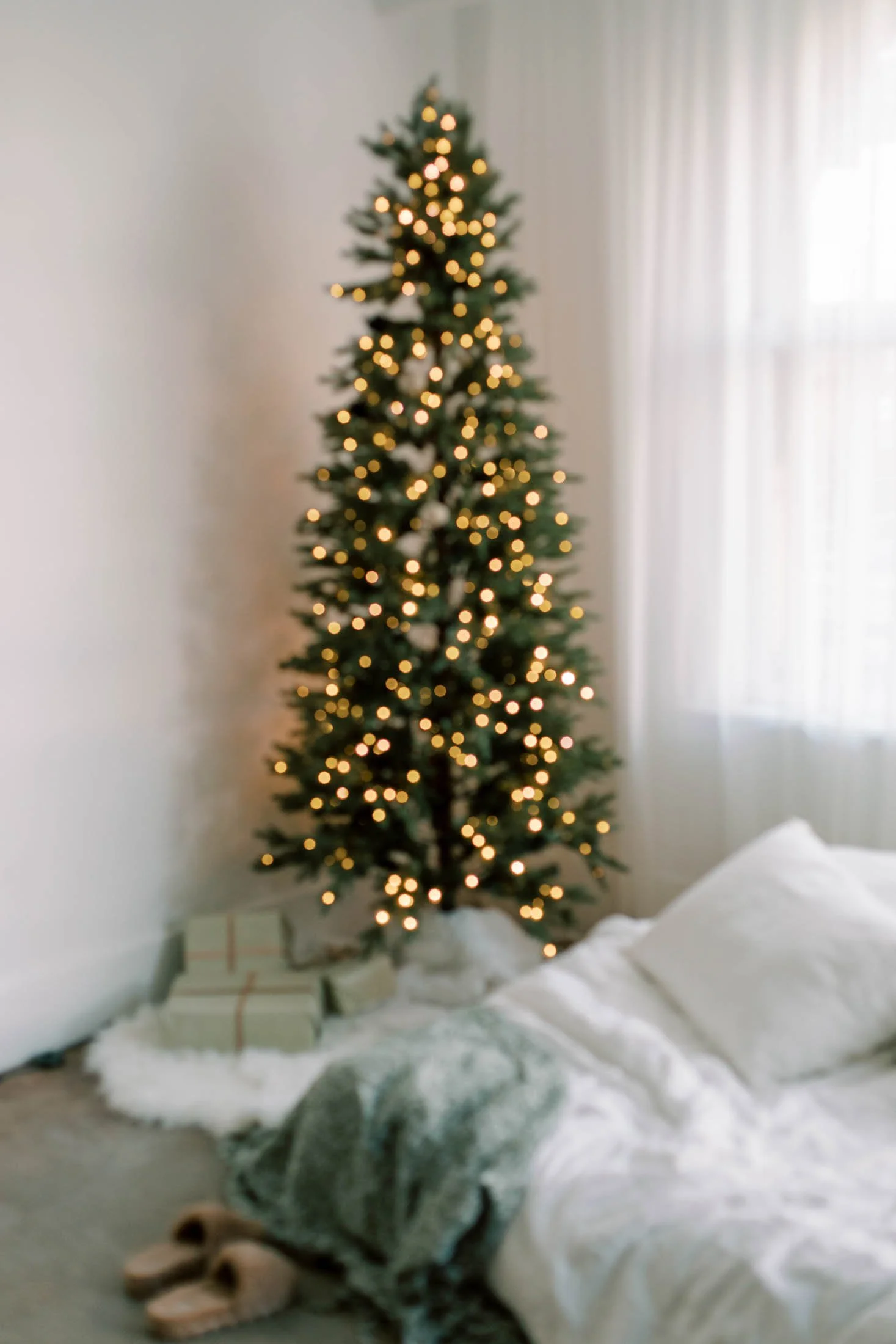 A decorated Christmas tree with lights in a room with light-colored curtains and a soft, fluffy white blanket or rug on the floor. There are wrapped presents at the base of the tree, and a gray tabby cat laying near the presents.