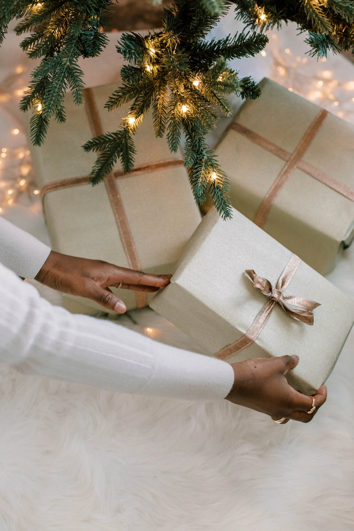 Decorative Christmas scene with hands holding a large beige gift box wrapped with a pinkish ribbon, placed under a Christmas tree with lit lights, on a soft white fur surface.