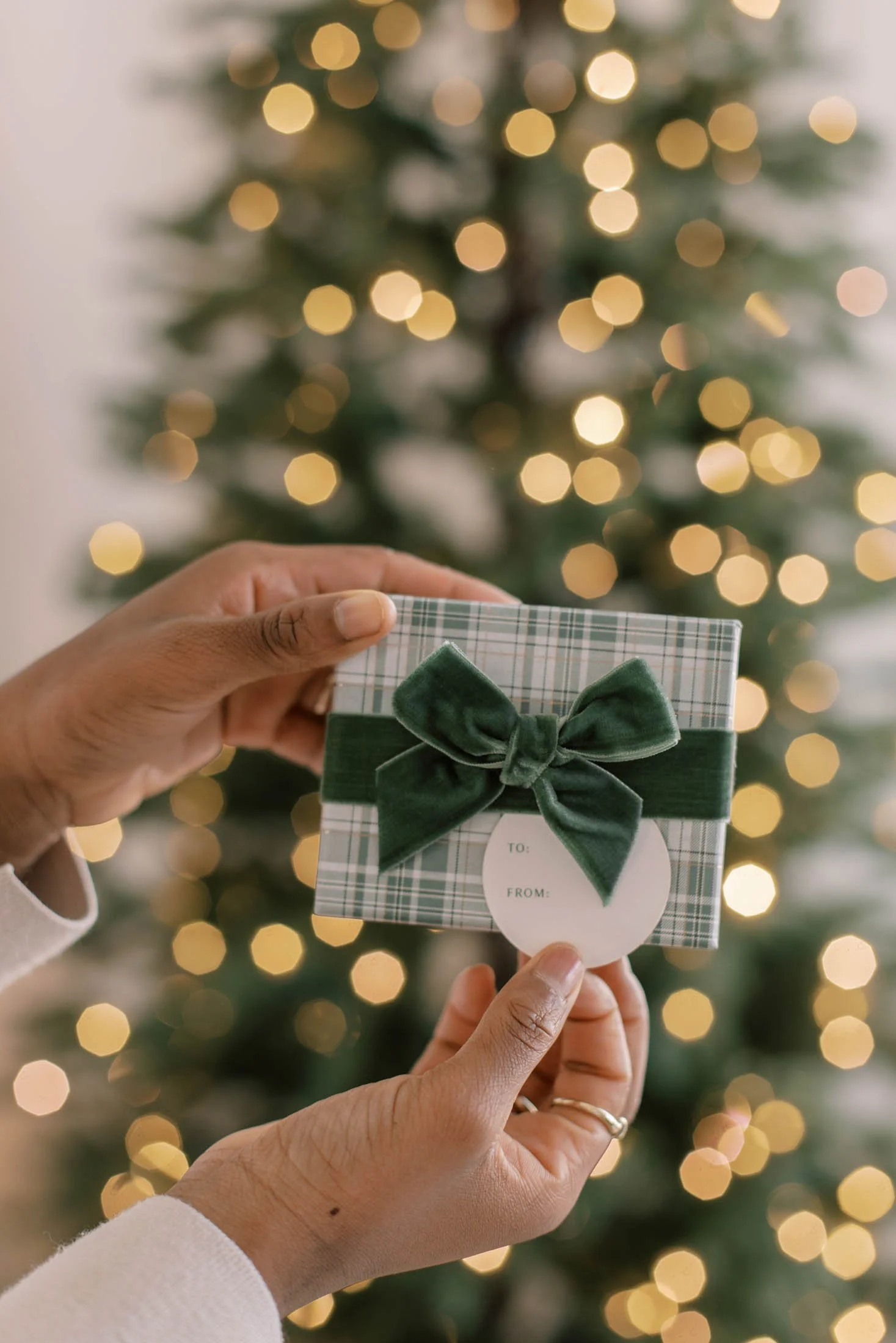 Person holding a Christmas gift box with a green velvet bow, in front of a decorated Christmas tree with blurred lights.