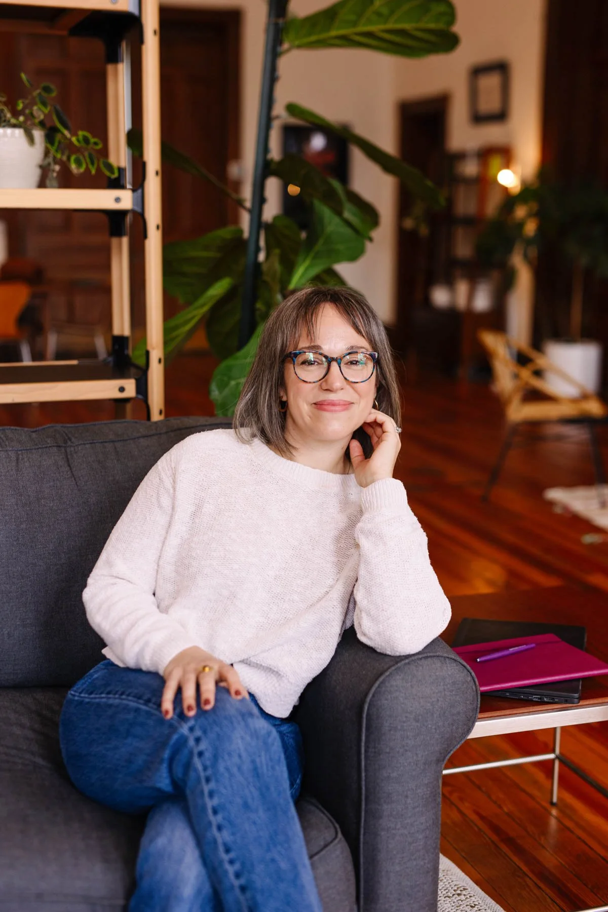 Woman with glasses smiling, sitting on a dark gray couch in a cozy living room, with a wooden floor, houseplant behind her, and a magazine and pen on a side table.