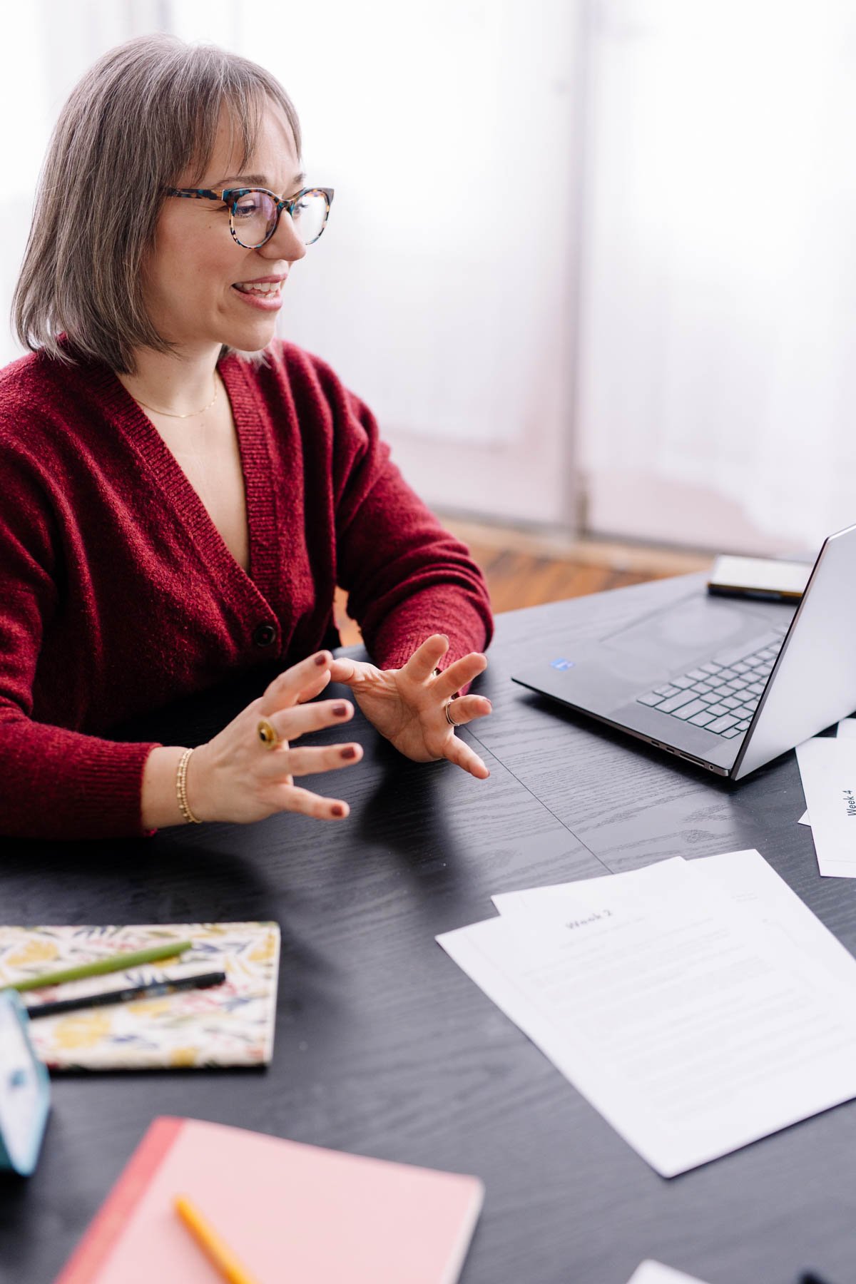 Woman in glasses and a red sweater sitting at a black desk with papers, a laptop, a notebook, and a colorful planner, engaged in conversation.
