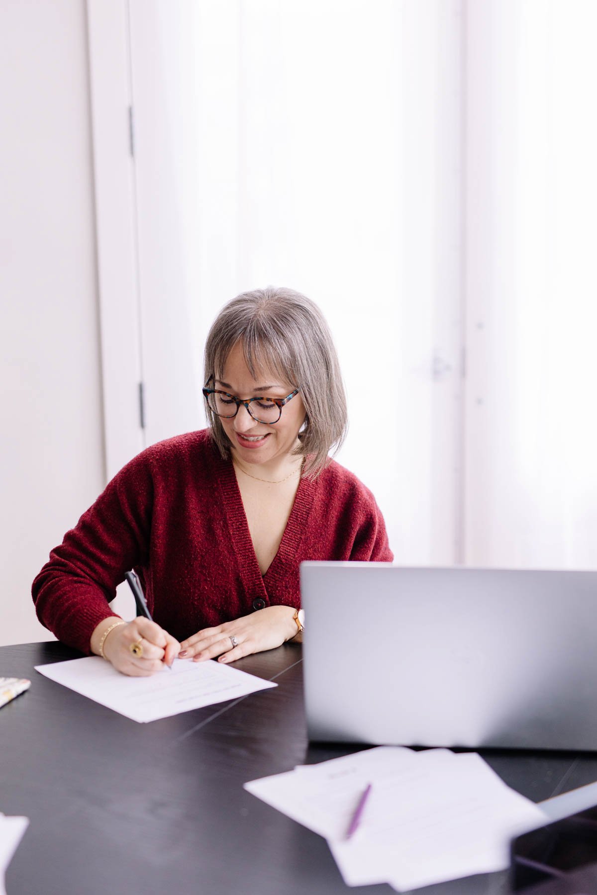 Woman with glasses and a red sweater sitting at a desk, writing on a piece of paper, with a laptop and notes on the desk.