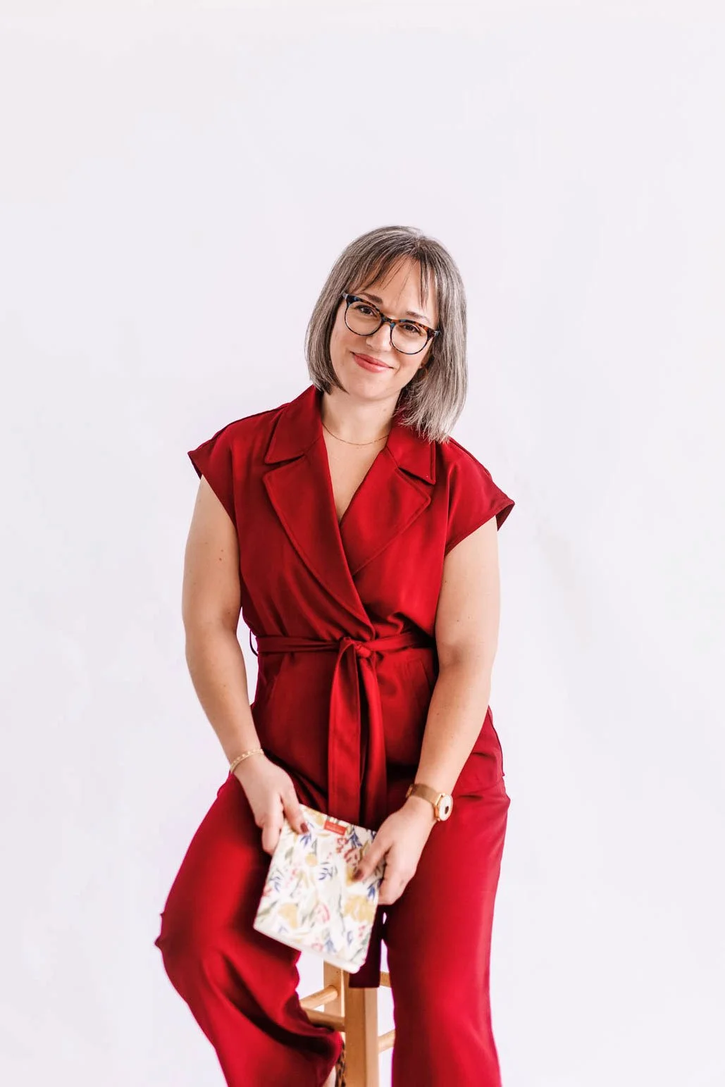 A woman with shoulder-length gray hair, wearing glasses and a red wrap dress, sitting on a stool against a white background, holding a floral-patterned clutch bag, smiling at the camera.