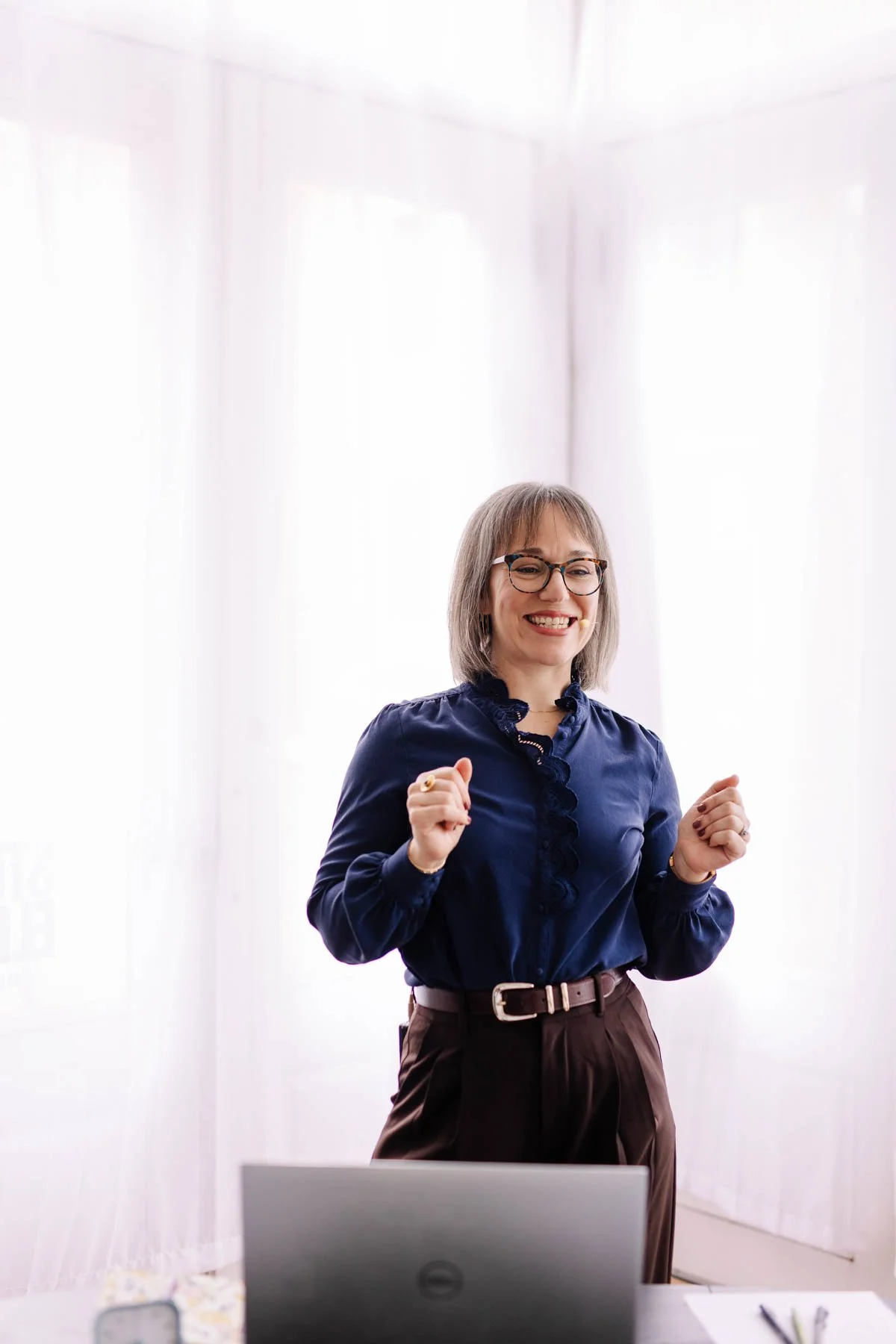 Happy woman with glasses wearing a navy blouse and brown pants, standing in an office with white curtains, smiling and showing a victory gesture with both fists.
