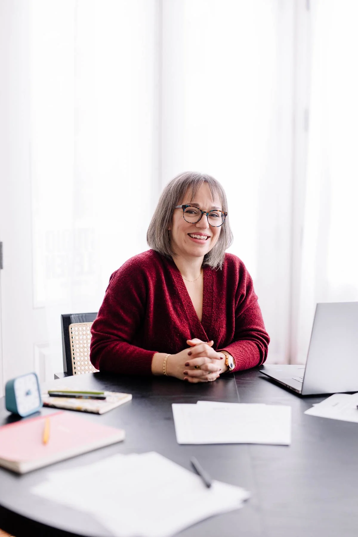A woman with gray hair and glasses, smiling, wearing a red sweater, sitting at a desk with a laptop, notebooks, and papers, in a bright office.