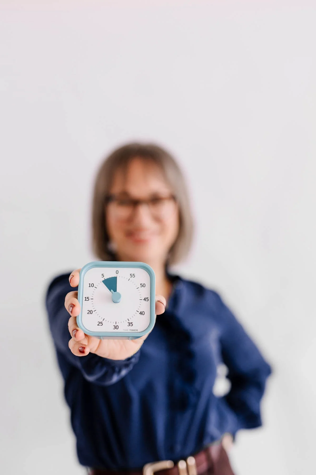 A woman holding a white and blue timer clock towards the camera, smiling and blurred in the background.