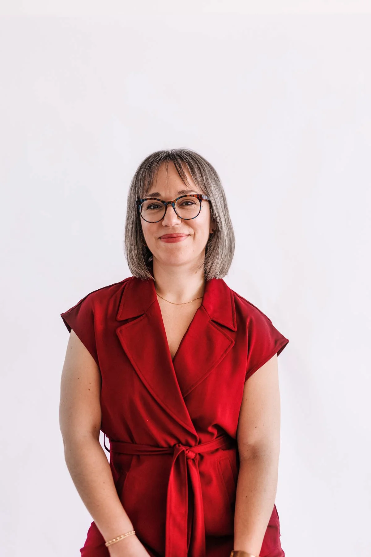 A smiling woman with shoulder-length gray hair, wearing glasses and a red sleeveless dress with a waist tie, standing against a white background.