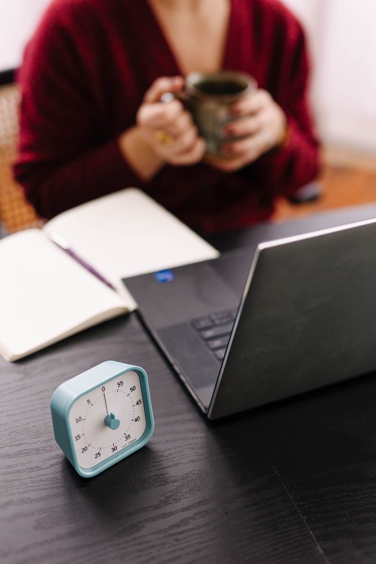 A person in a red top holding a coffee mug in front of a laptop, with an open notebook, a small clock, and a timer on a dark wooden table.
