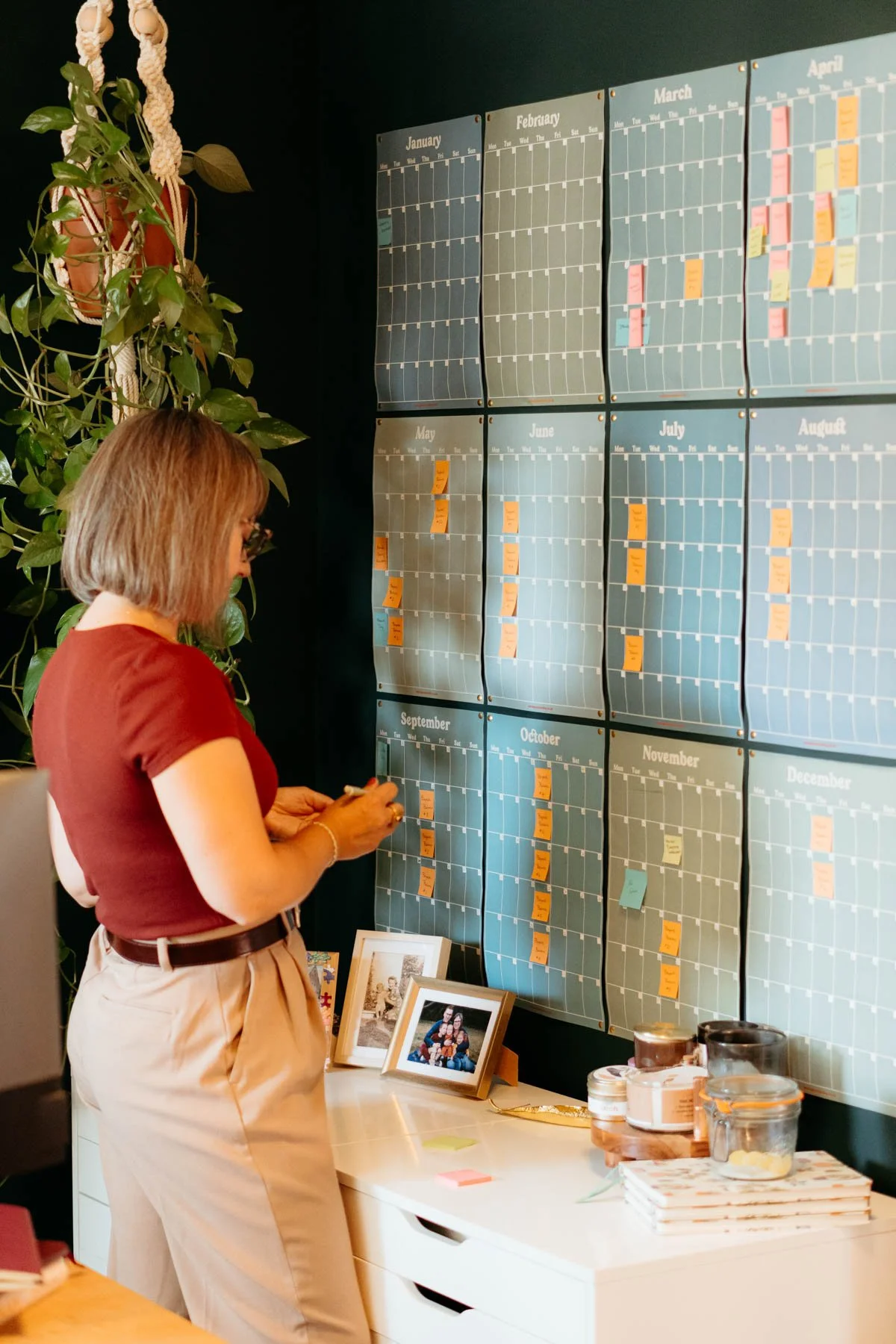 A woman in a brown shirt and beige pants organizing sticky notes on a large wall calendar displaying months from January to December. The calendar has multiple colored notes, and there are framed photos and various jars on a white table below.