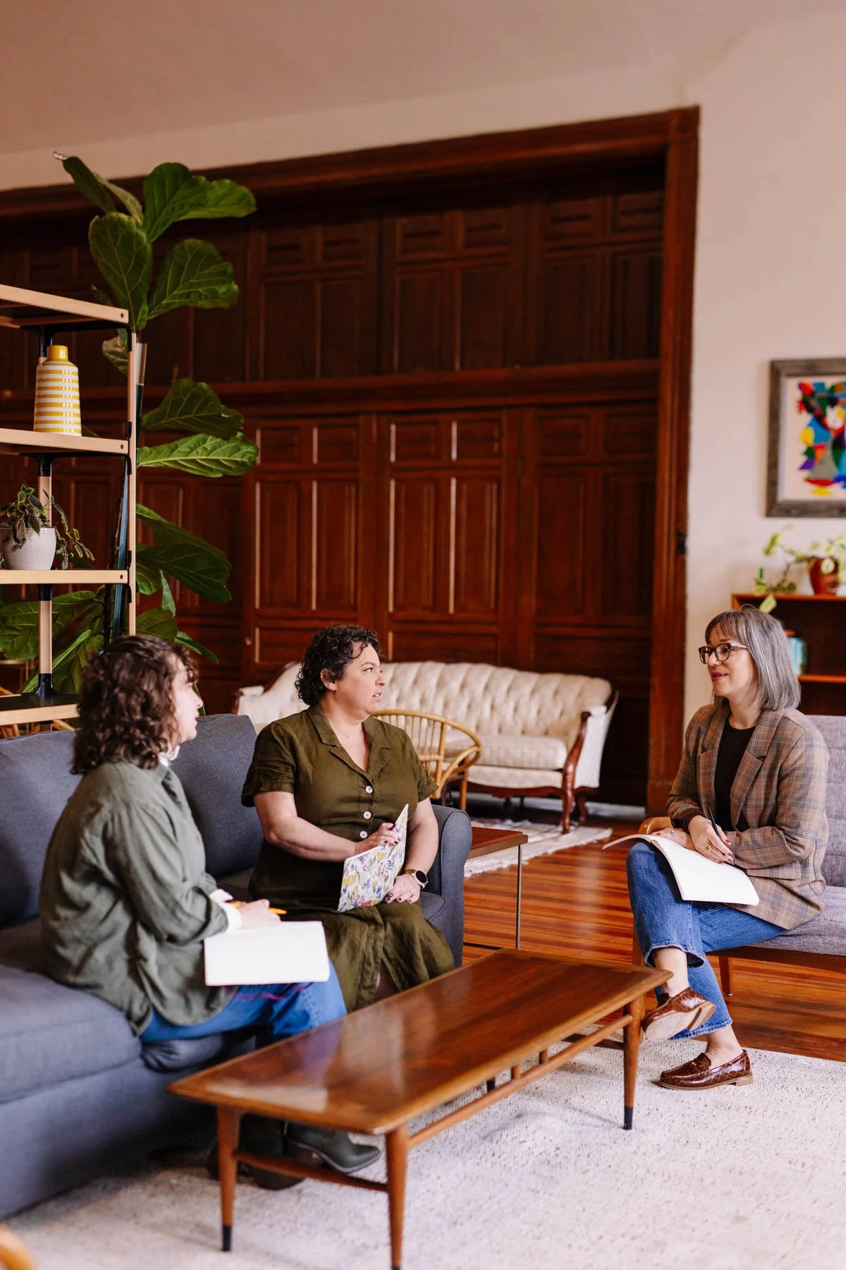 Four women seated in a living room having a conversation. Three women are sitting on a couch, one is sitting on a chair with a notebook.