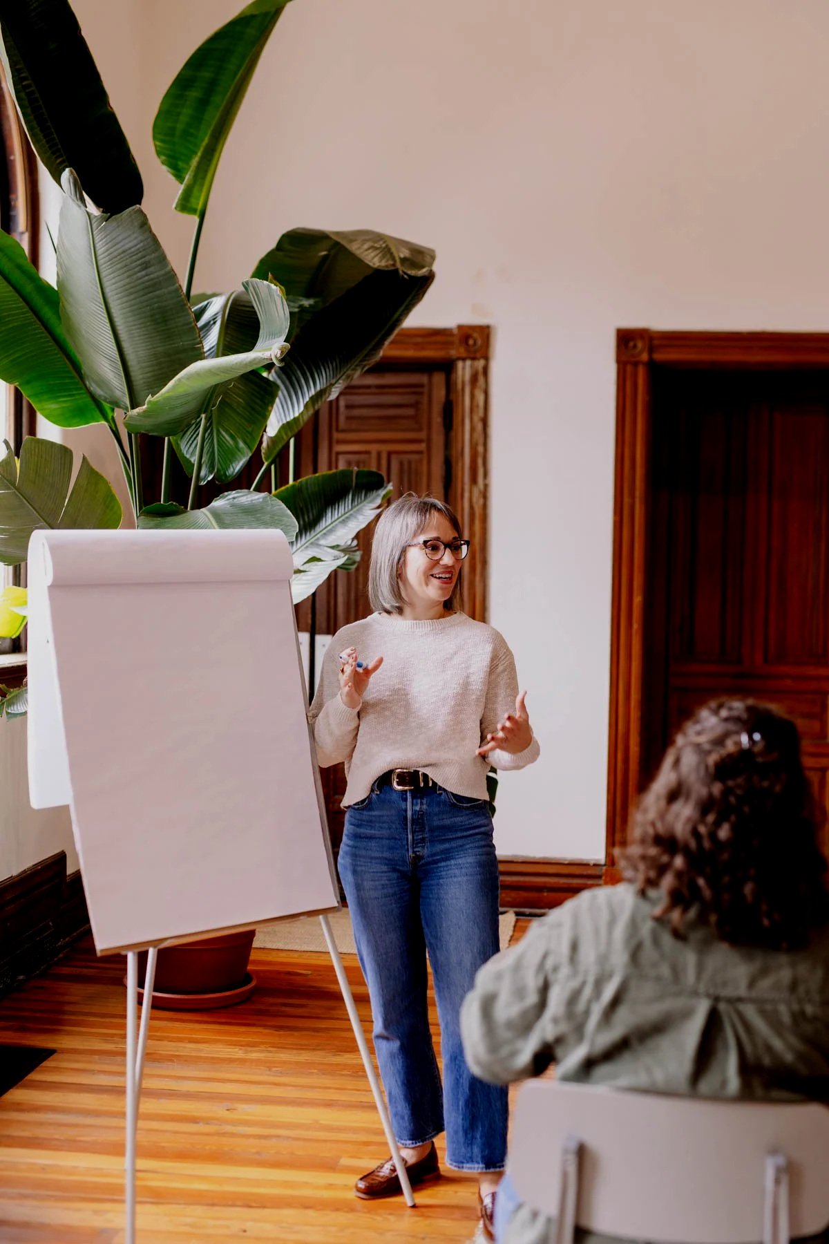 A woman with glasses, wearing a beige sweater and blue jeans, stands in front of a whiteboard, smiling and gesturing with her hands as she teaches or presents to an audience in a room with wooden flooring, large green plant, and wooden window frames.