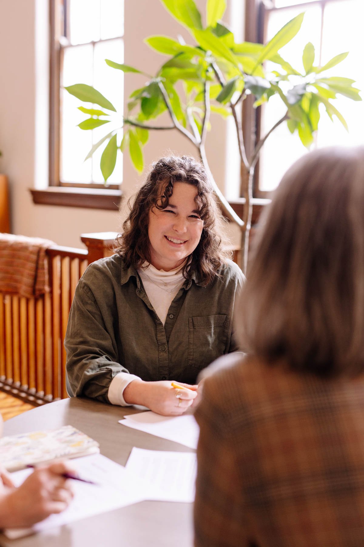A woman smiling and talking to another person at a table indoors with large windows and a potted plant in the background.