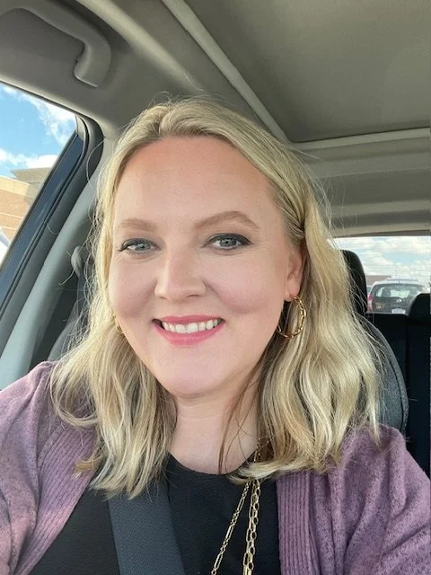 A smiling woman with blonde hair and gold hoop earrings, sitting in a car during daytime.