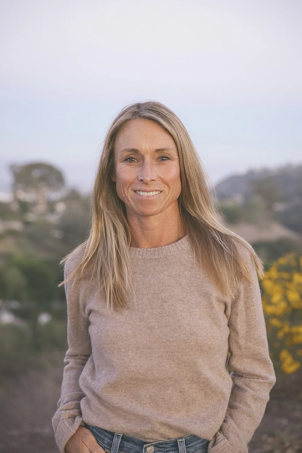 A woman smiling outdoors with a hillside and trees in the background during daylight.