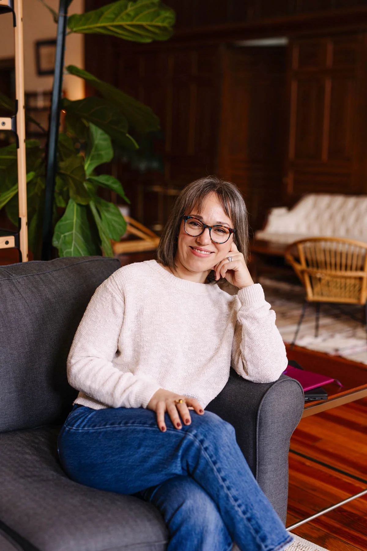 A woman with glasses and brown hair sitting on a dark gray sofa, smiling and resting her chin on her hand in a cozy, well-lit room with wooden furniture and a large leafy plant behind her.