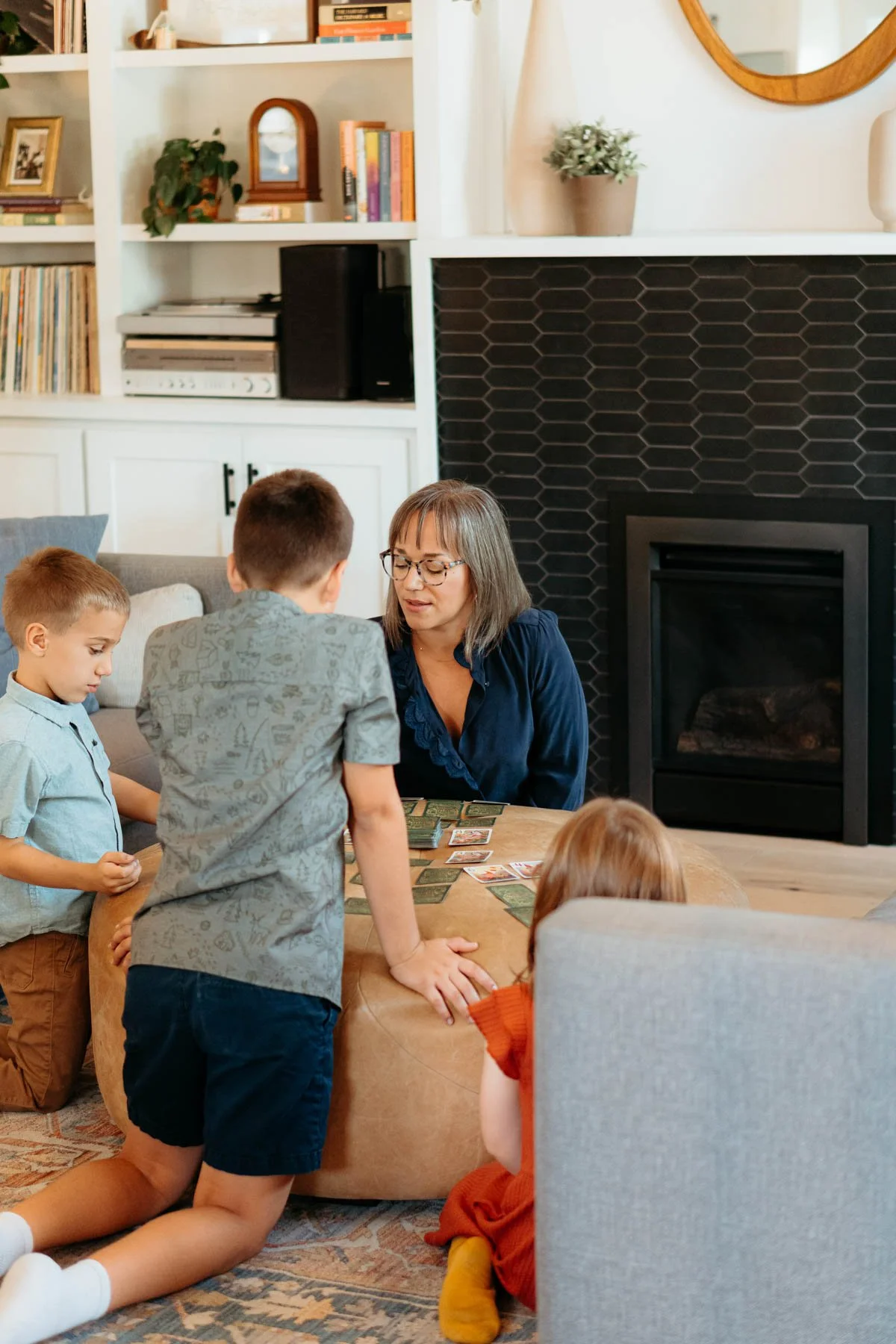 A woman playing a card game with four children around a wooden table in a living room with a black fireplace, white bookshelf, and wall decor.