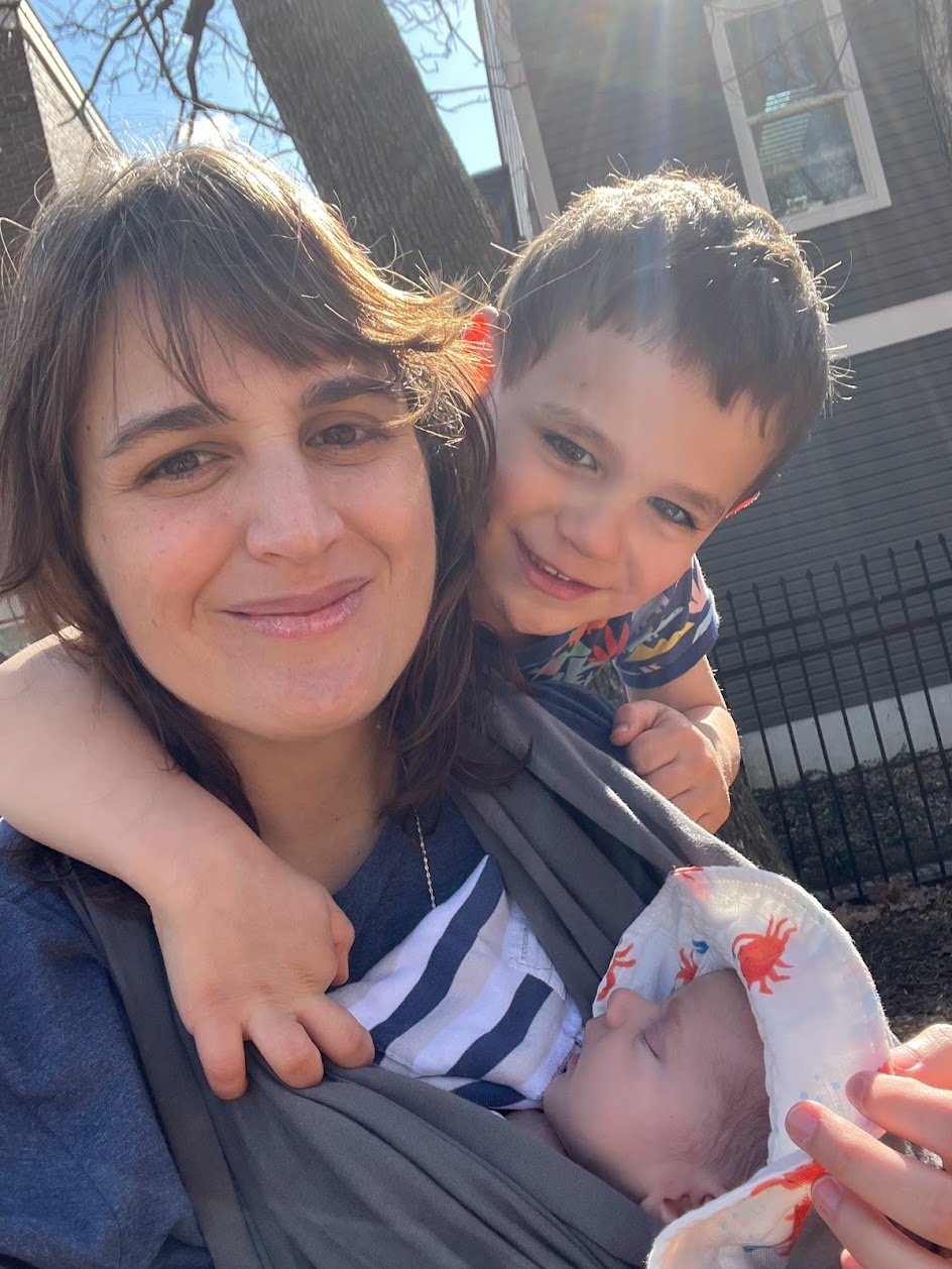 A woman smiling and carrying a baby in a baby carrier, with a young boy hugging her from behind. The baby is sleeping and wearing a hood with orange crabs. The sun is shining, and a house with a window and a tree are visible in the background.