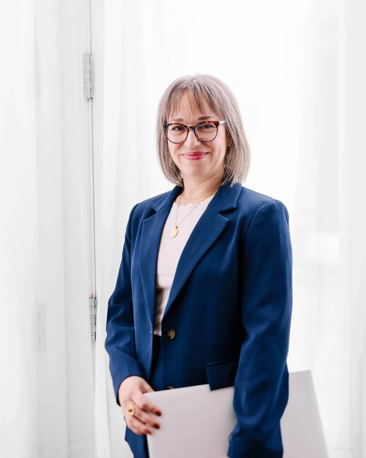 A professional woman with short, light brown hair and glasses, wearing a navy blue blazer and white top, holding a white folder, standing in front of a white background.