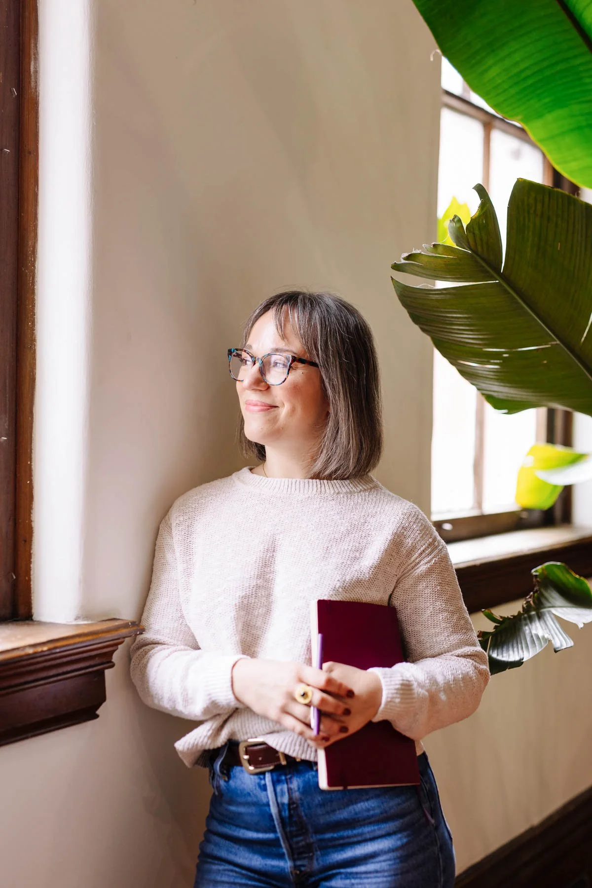 A woman with glasses and shoulder-length hair stands by a window, holding a maroon notebook and a purple pen, smiling softly, near large green plants inside a room with wooden trim.