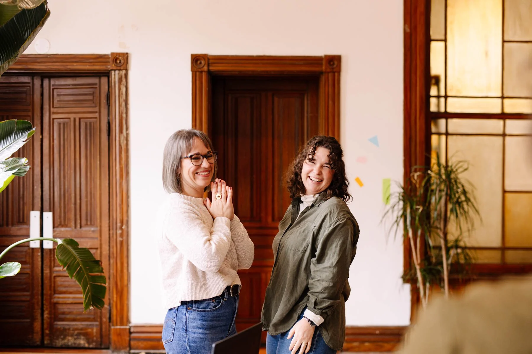 Two women smiling and laughing indoors, standing near a white wall with wooden accents and a large window, surrounded by indoor plants.