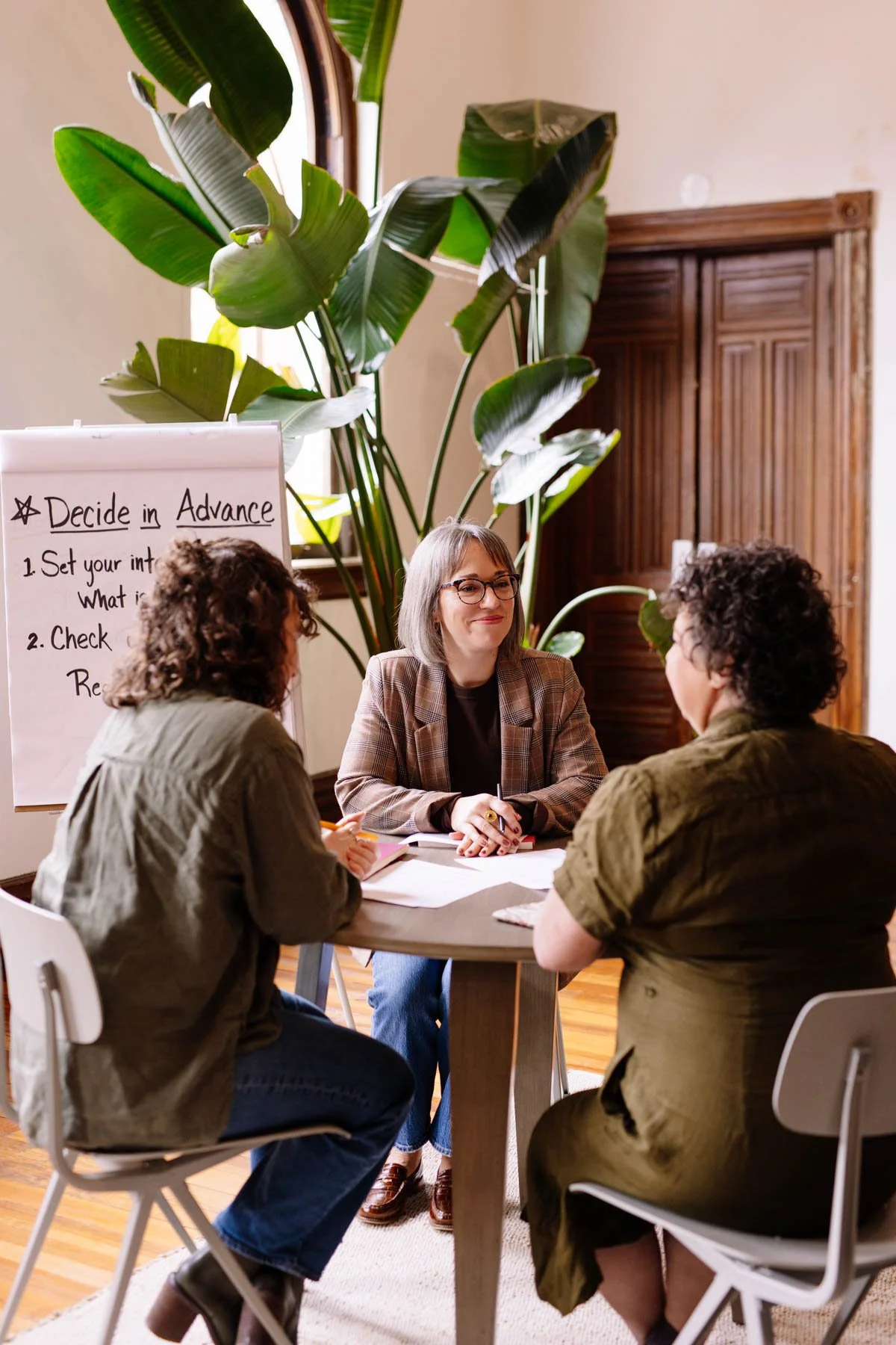 Three women having a discussion at a table in a room with large green indoor plants and a whiteboard with notes. One woman is wearing glasses and a plaid blazer, while the other two women are listening.