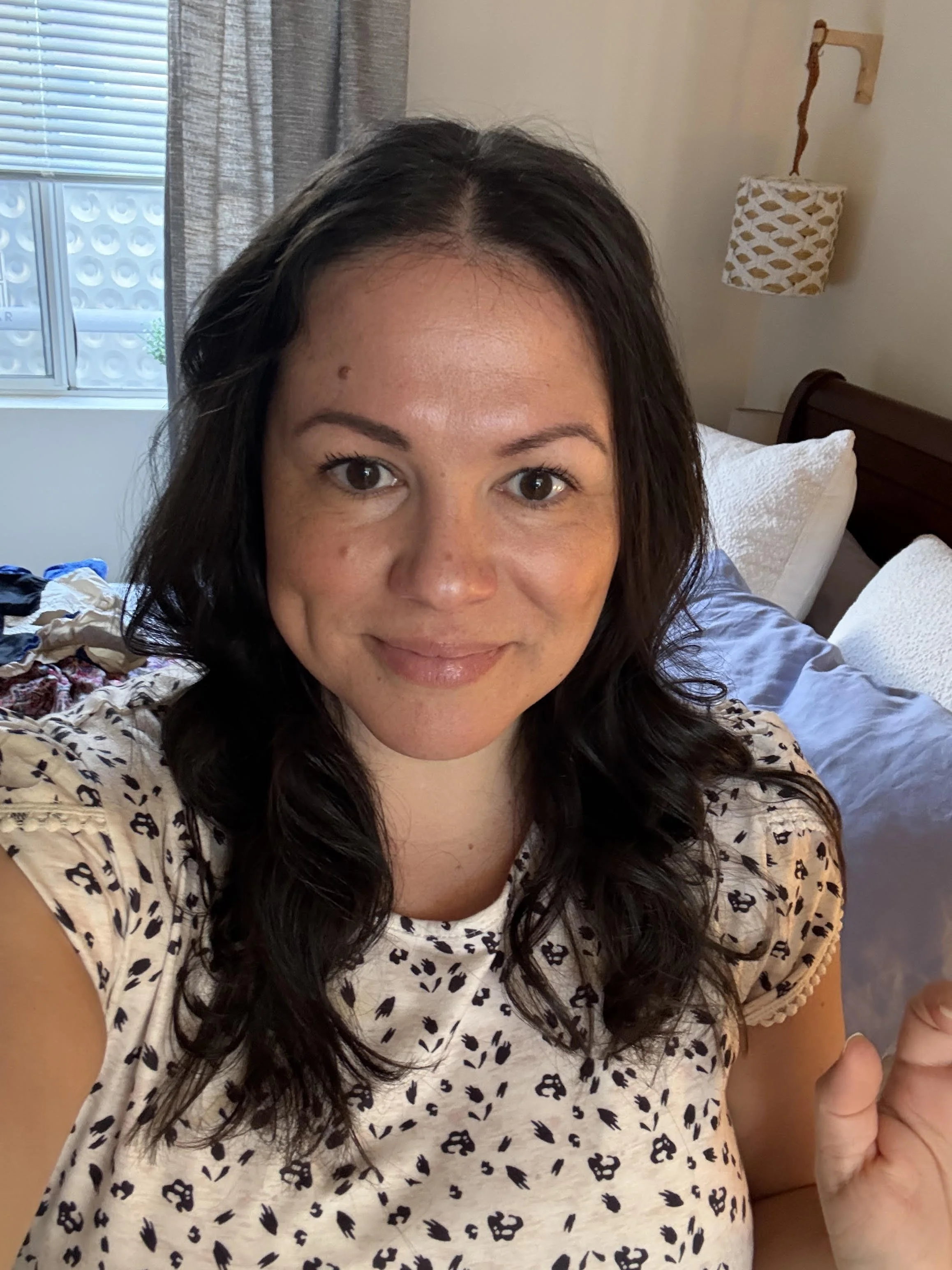 Close-up of a smiling woman with dark brown curly hair, wearing a white shirt with black animal print, sitting on a bed in a room with curtains and a window in the background.