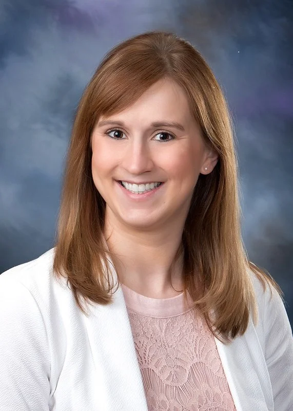 A young woman with shoulder-length light brown hair, smiling, wearing a pink lace top and white blazer against a dark, cloudy background.