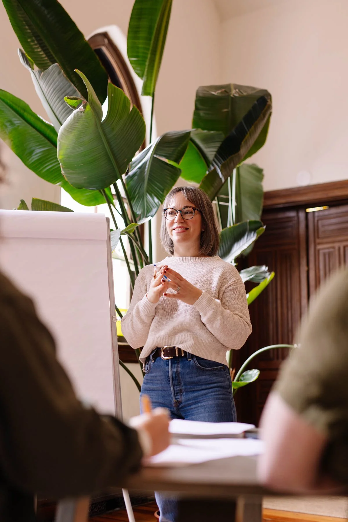 A woman wearing glasses, a beige sweater, and jeans stands in front of a large, leafy plant, smiling and clasping her hands together during a presentation or lecture.