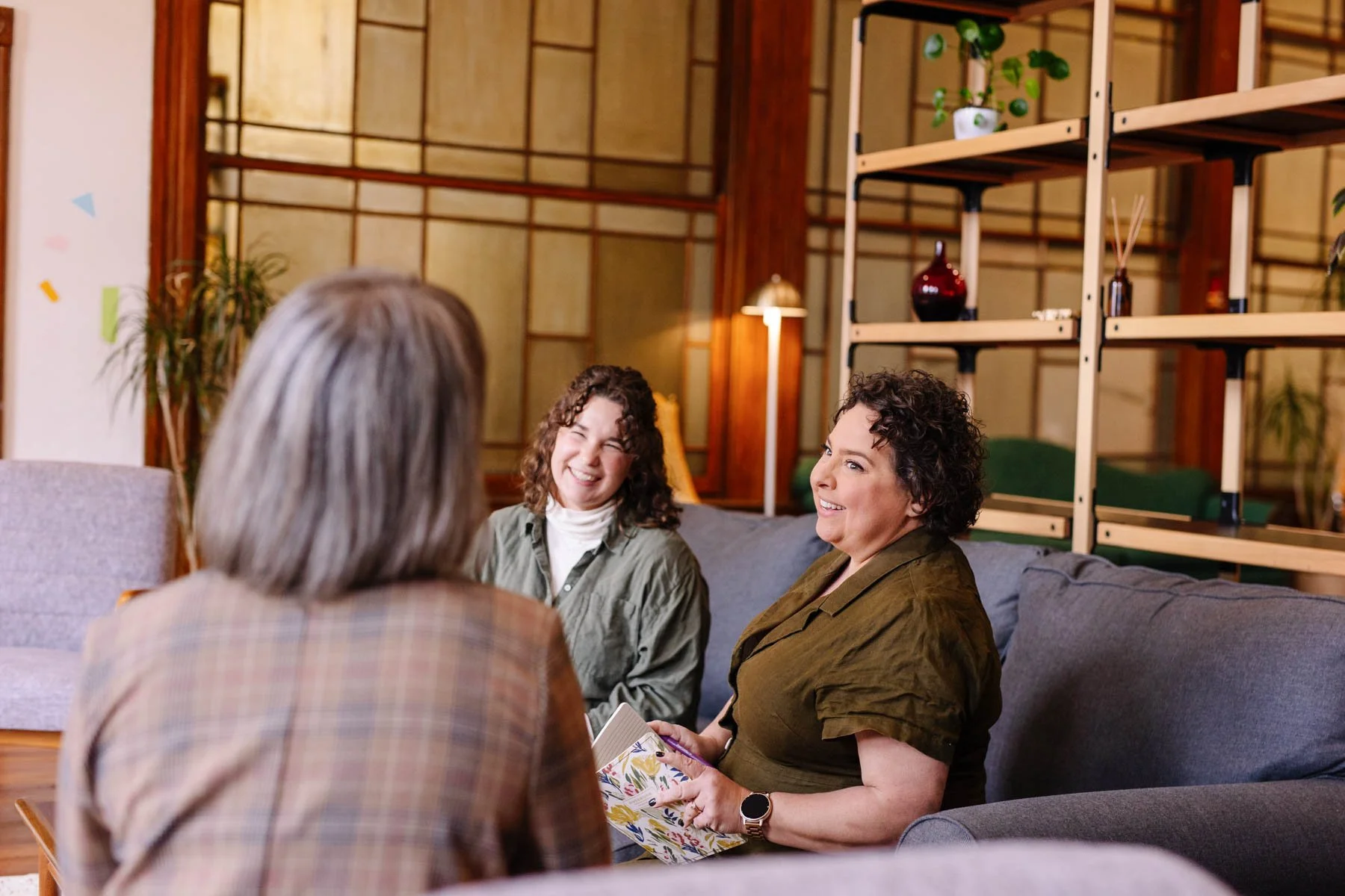 Four women sitting on a sofa and engaging in conversation in a cozy living room with wooden accents and decorative shelves.