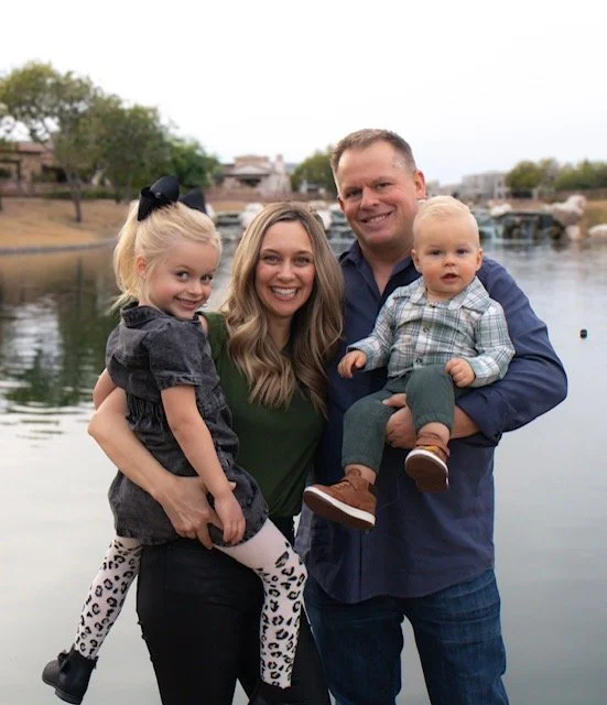 Family of four outdoors by a body of water, smiling at the camera. The mother holds a young girl, and the father holds a young boy.