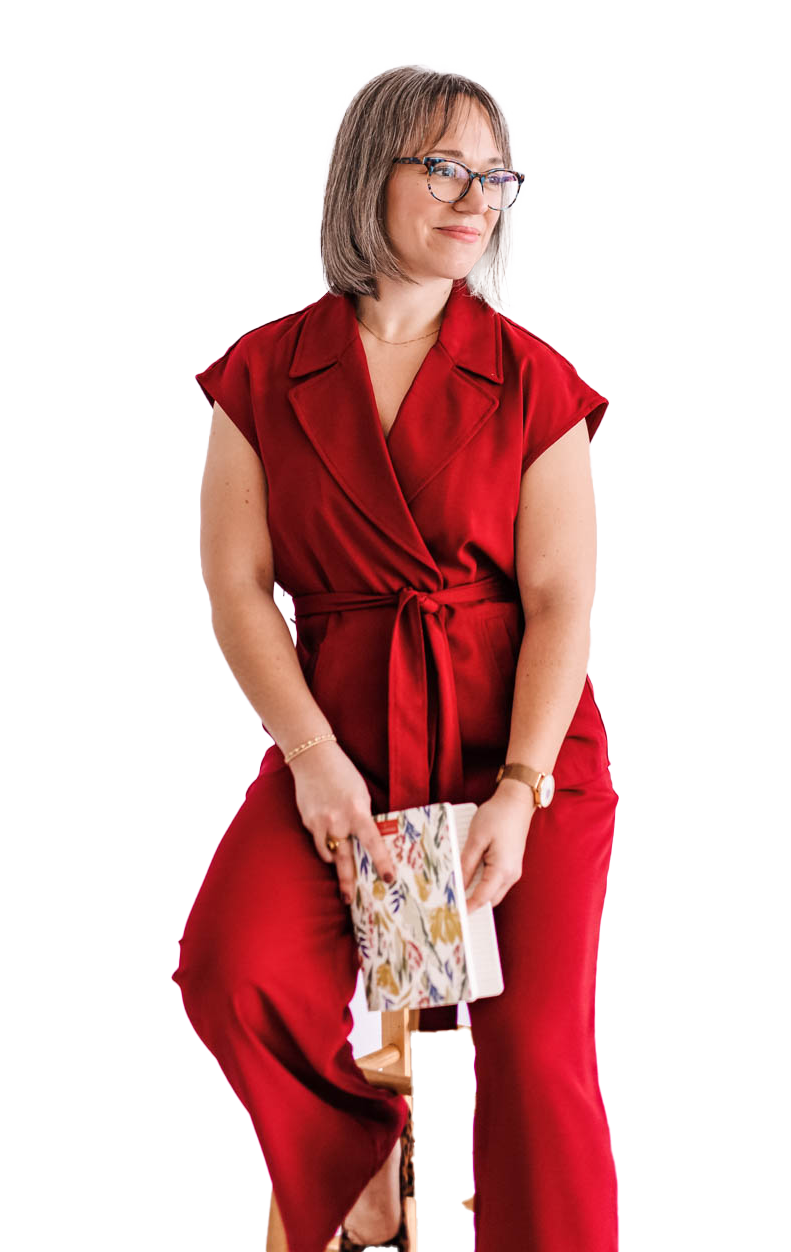 A woman with shoulder-length gray hair and glasses, wearing a red top and red pants, sitting on a wooden stool and holding a floral-patterned notebook.