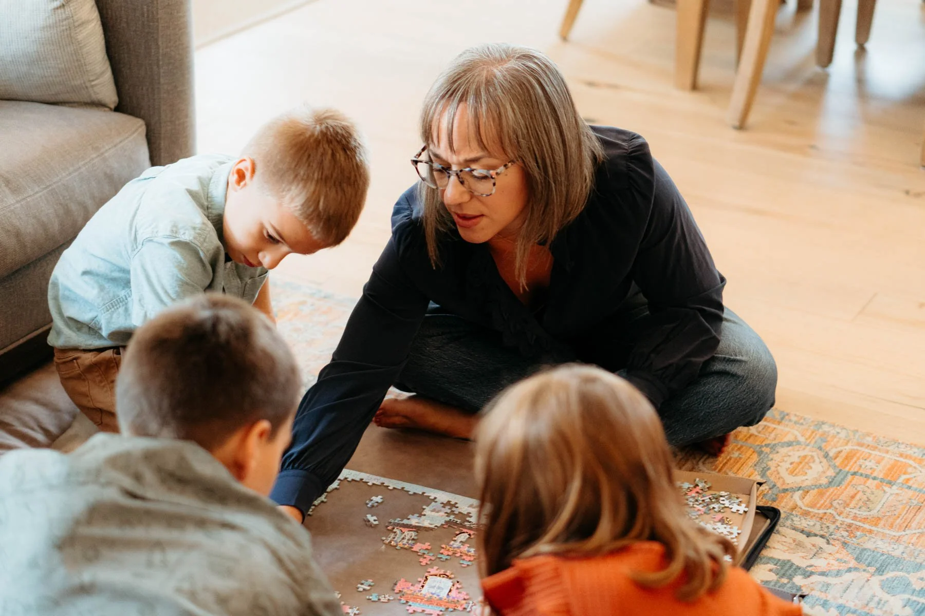 A woman with glasses and shoulder-length brown hair is sitting on the floor with children, working on a jigsaw puzzle. The kids are sitting around her, all focused on the puzzle pieces in front of them. The room has wooden floors, a beige rug, and a dining table in the background.