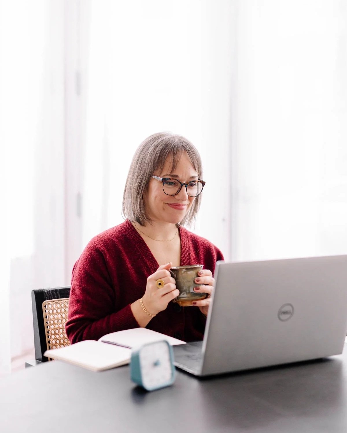 A woman with glasses and shoulder-length gray hair, wearing a red sweater, sits at a desk holding a mug, in front of an open laptop, with a notebook nearby.