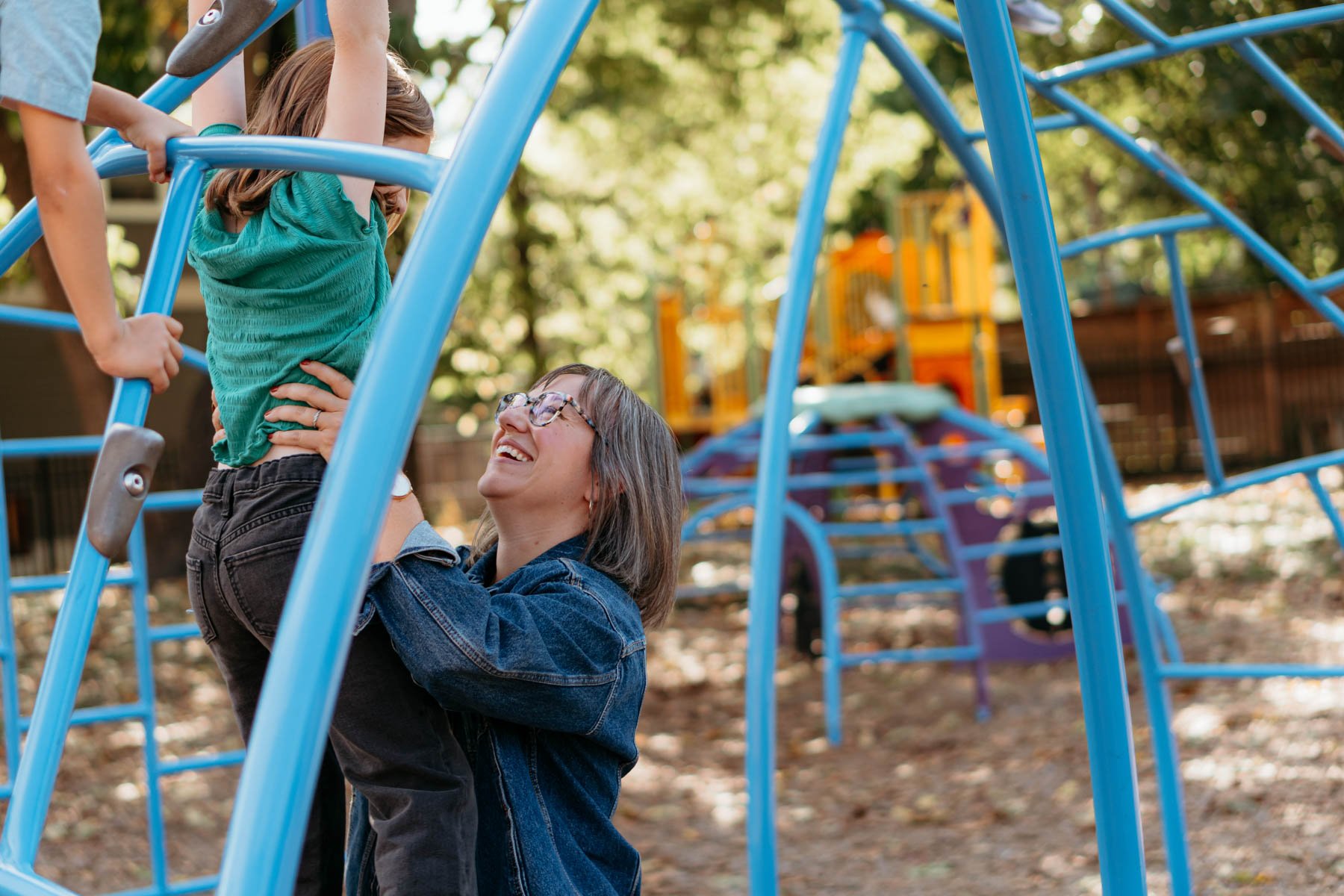 A woman and a young girl are playing on a blue jungle gym in a park. The woman is holding the girl up, and they are smiling at each other. There are trees and a colorful playground structure in the background.