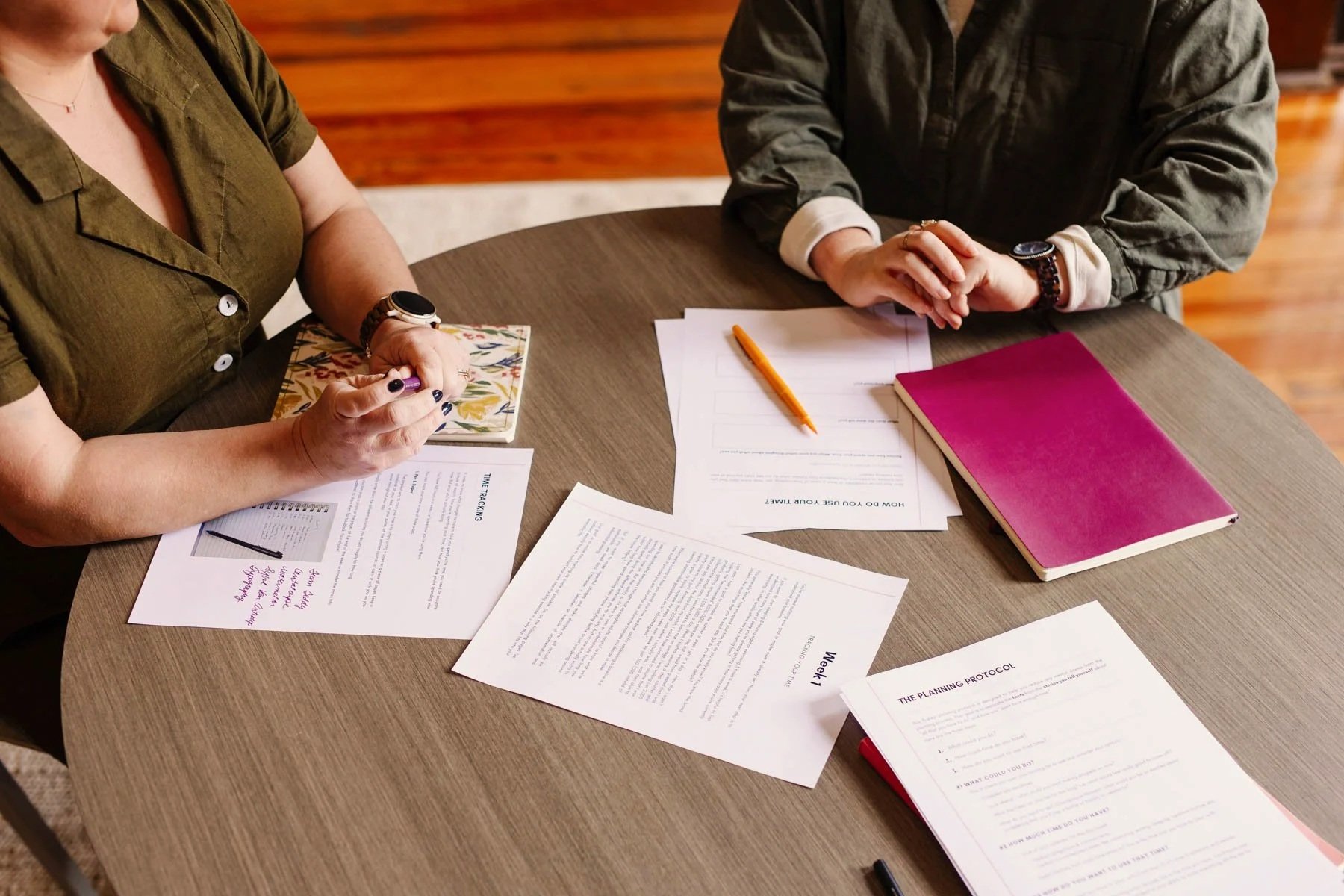 Two people sitting at a round wooden table with various papers, a purple notebook, and an orange pen. One person is holding a pen and a notebook, and the other has their hands clasped on the table. The setting appears to be a workspace or meeting area.