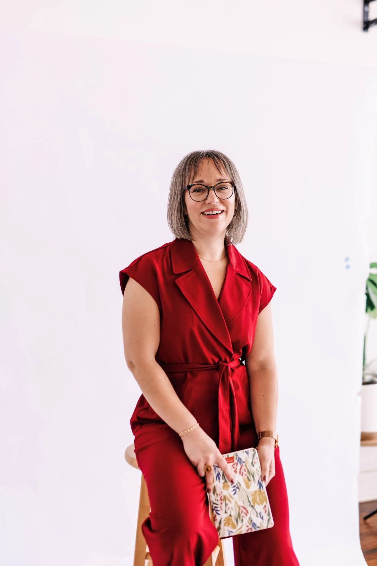 A smiling woman with glasses and short gray hair, wearing a red sleeveless dress tied at the waist, sitting on a stool with a floral notebook in her lap, in a minimal white room with a small plant visible in the background.