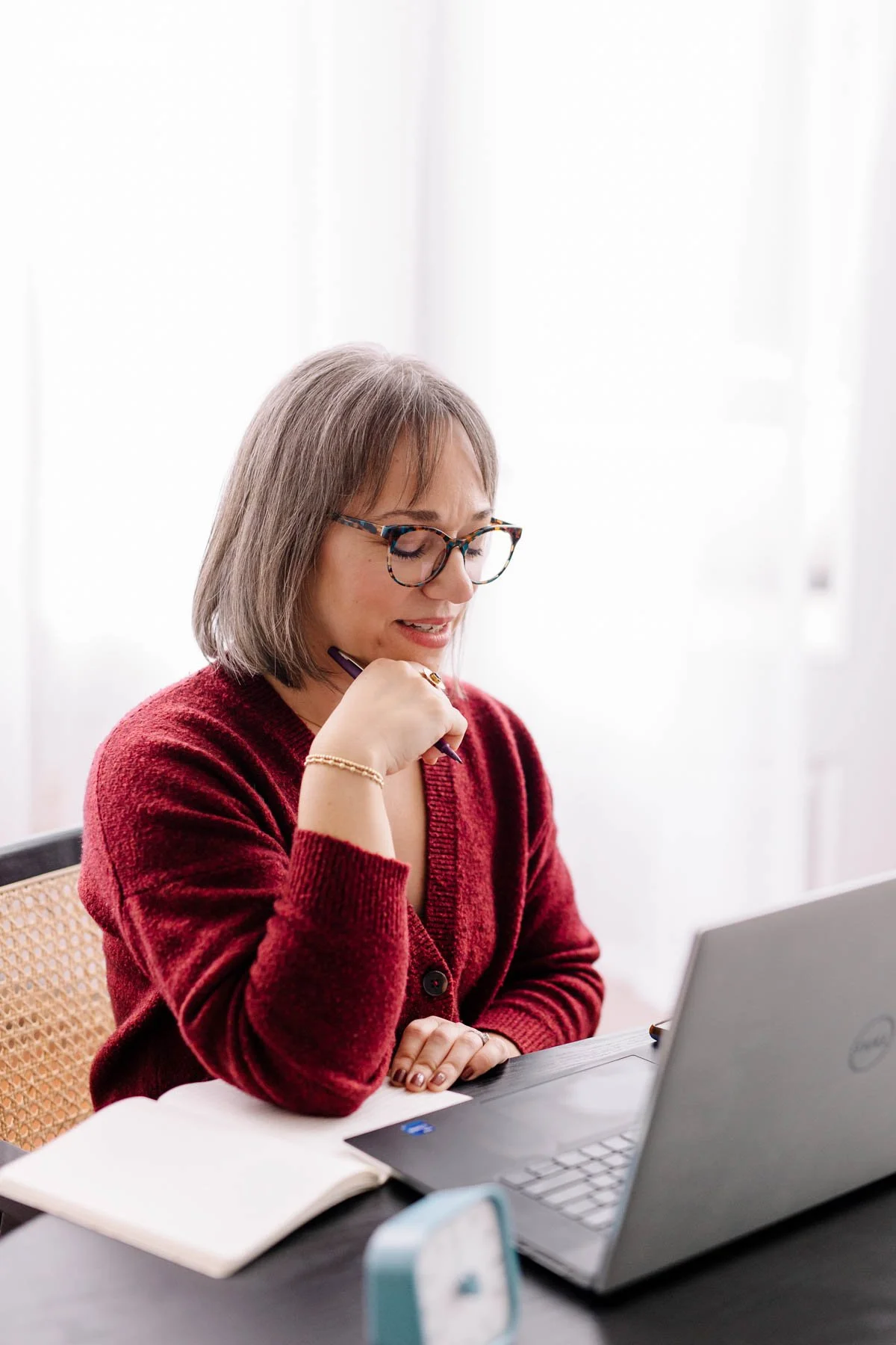 A woman wearing glasses and a red cardigan is sitting at a desk with a laptop, holding a pen near her face, looking down at the laptop screen.