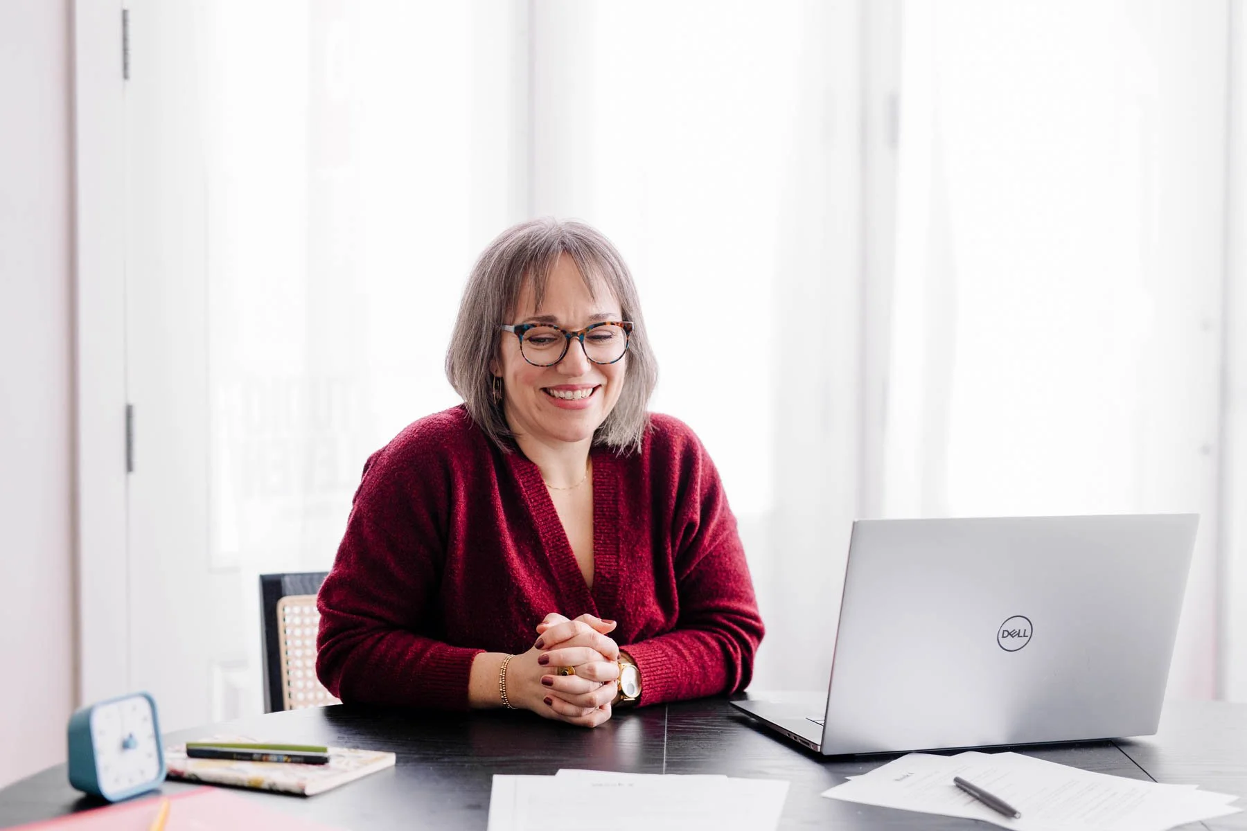 A woman with gray hair, wearing glasses and a red sweater, sitting at a black desk with a Dell laptop, smiling with her hands clasped, in a bright office with white walls and large windows.