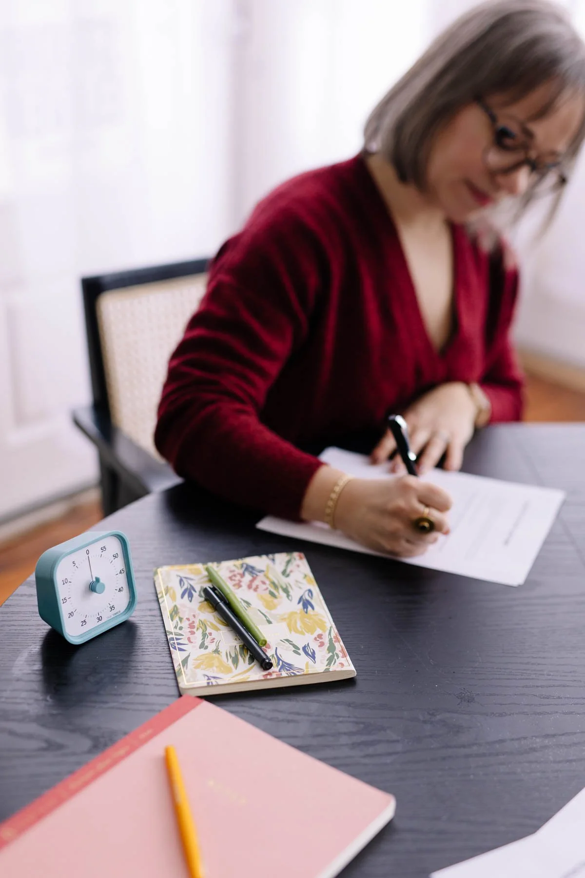 A woman with glasses in a red top is sitting at a table, writing on a sheet of paper with a black pen. On the table, there is a pink notebook with a yellow pencil, a floral notebook with two pens, a blue timer, and some papers.