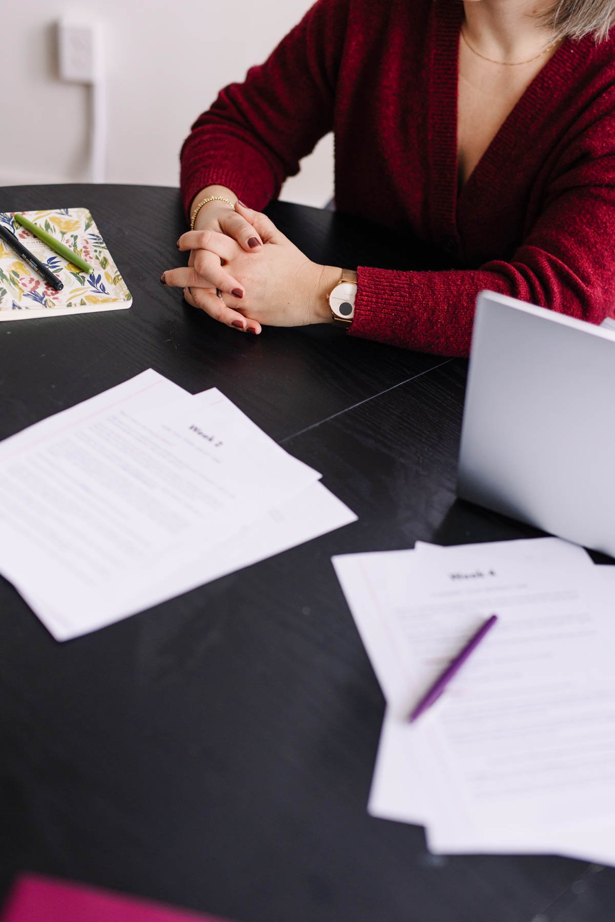 A woman sitting at a desk with papers, pens, and a laptop, wearing a red sweater and bracelet, with her hands clasped.