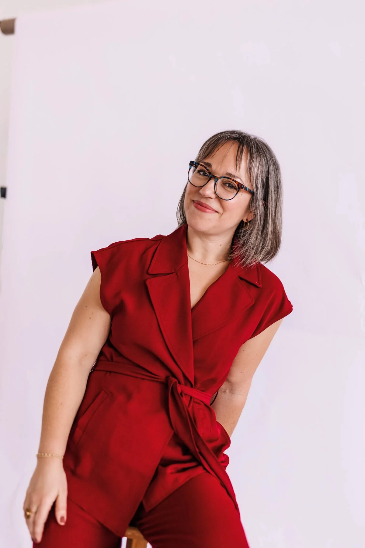 Woman wearing glasses and a red sleeveless jumpsuit, posing against a plain white background.