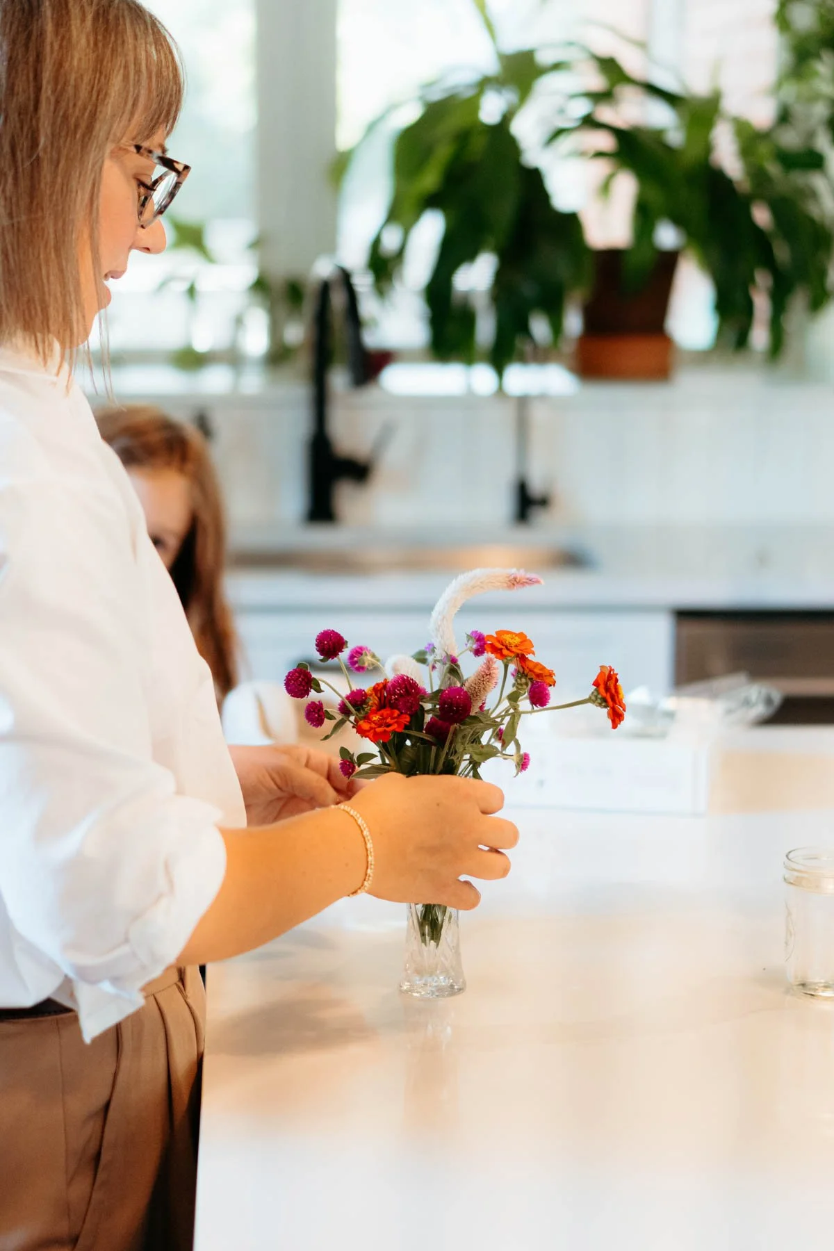 A woman with glasses arranging colorful flowers, including orange and purple blooms, in a clear glass vase on a white countertop in a bright kitchen.