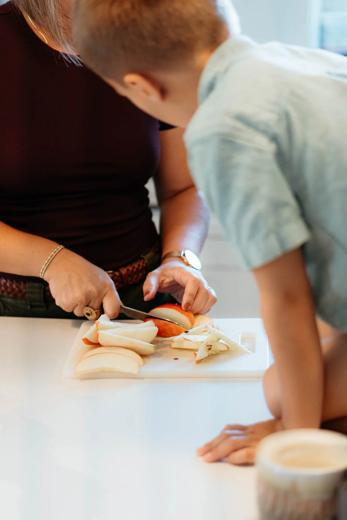 A woman and a young boy slicing apples on a white cutting board in a kitchen.