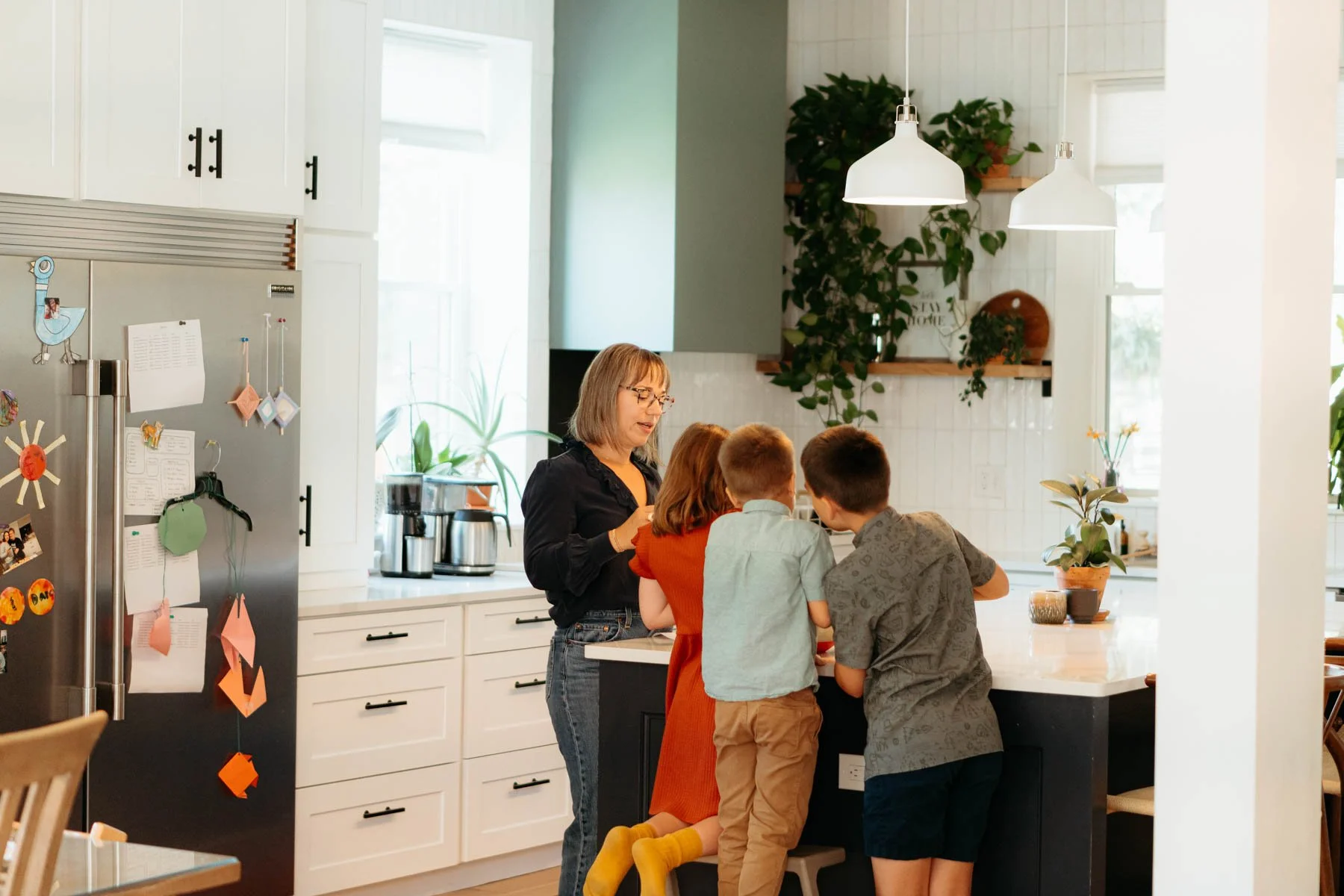 A woman and four children gathered around a kitchen island, with the woman interacting with one of the children, in a modern kitchen with white cabinets, hanging lights, and green plants.