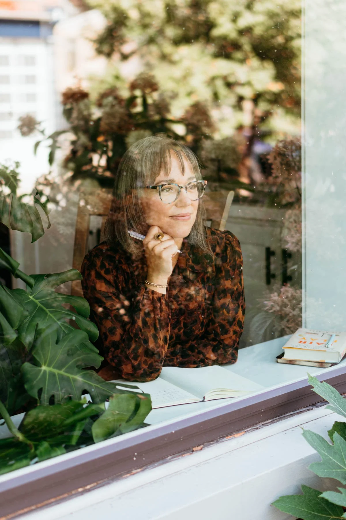 A woman with glasses and brown hair sits at a table, seen through a window with plants and a book on the table.