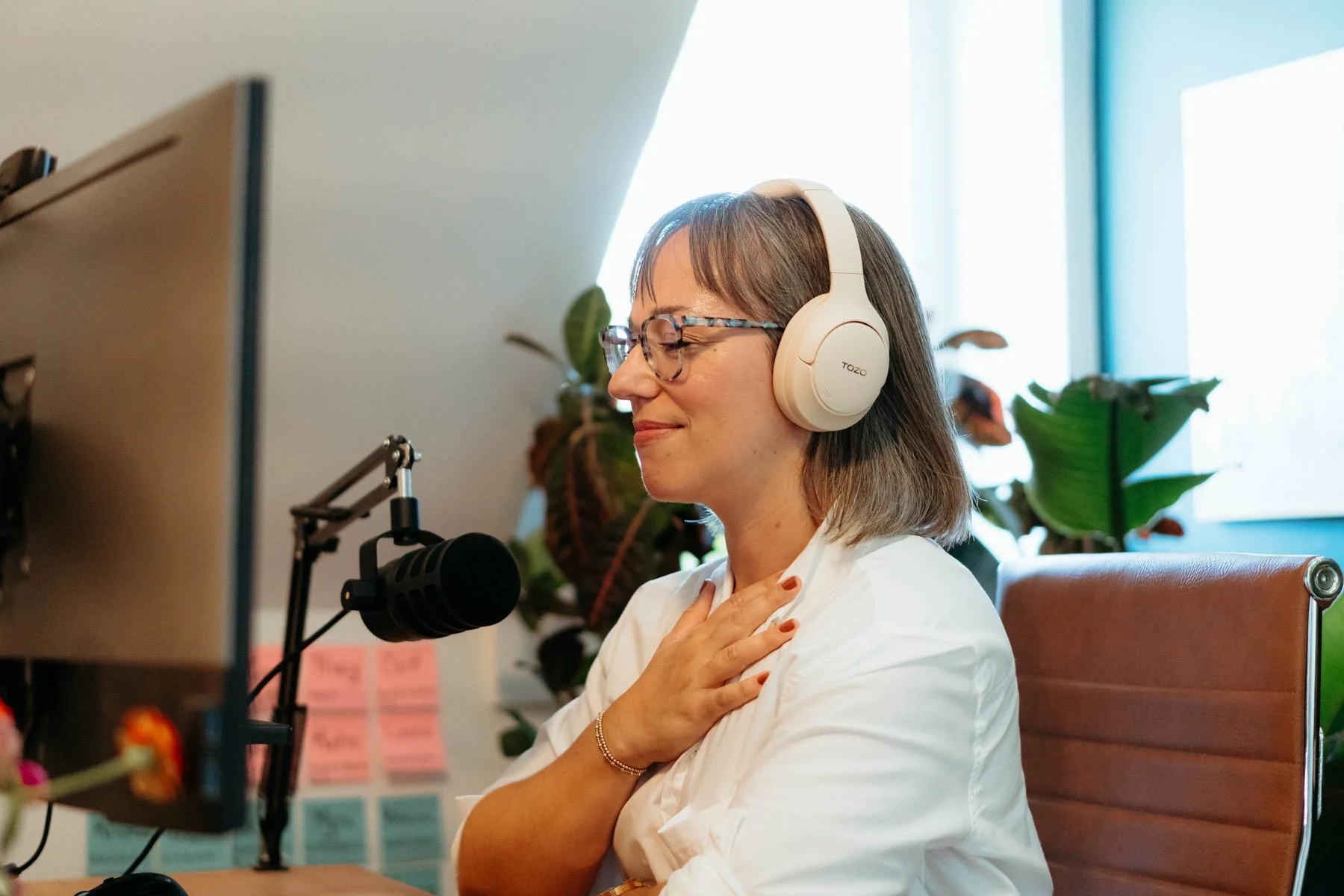 Woman with glasses and a white blouse, sitting at a desk with a microphone, listening with her hand on her chest in a bright office with plants and a computer monitor.
